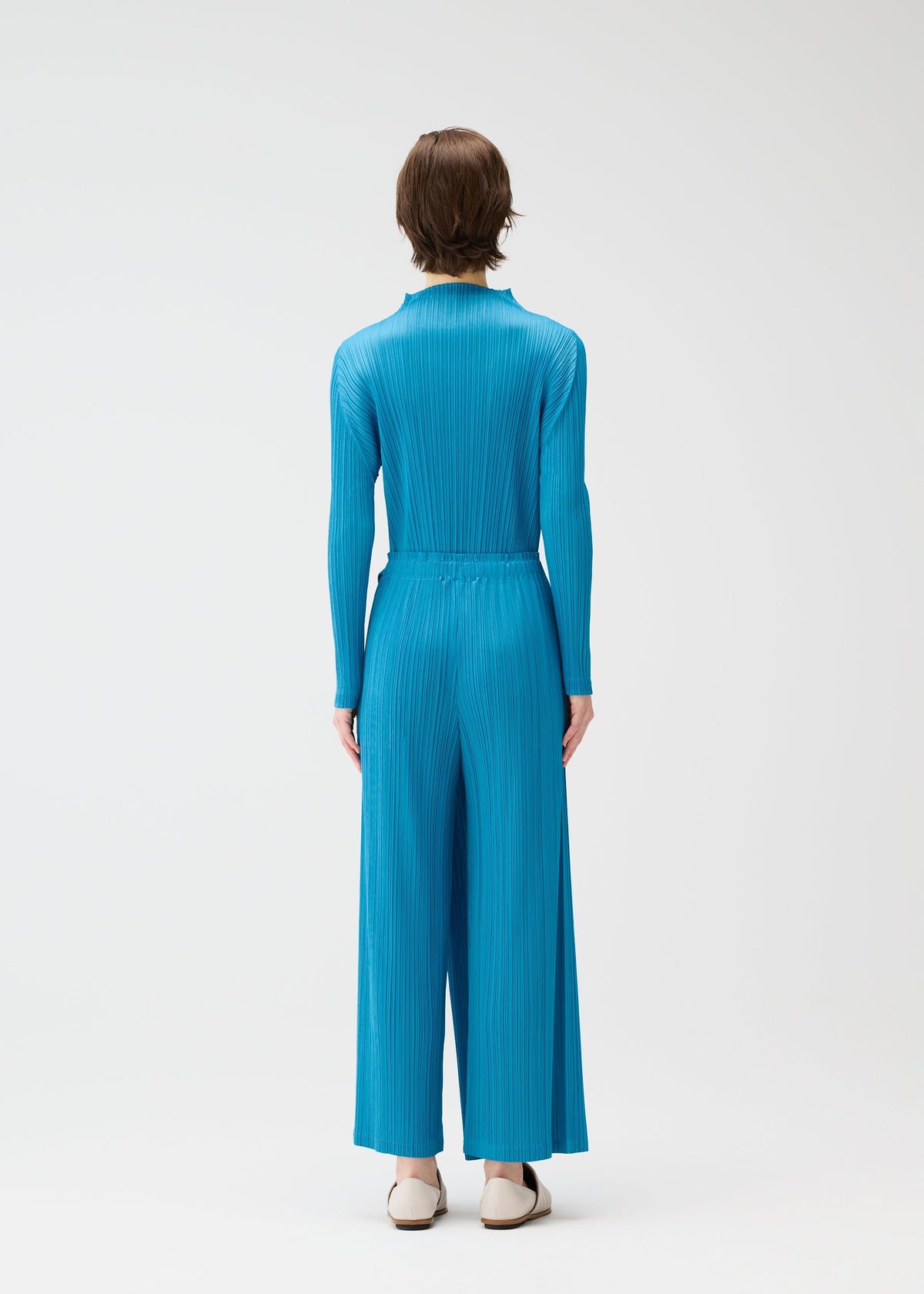 A person with short brown hair stands with their back to the camera, wearing the FACADE pleated jumpsuit by PLEATS PLEASE ISSEY MIYAKE and white shoes, set against a plain white background. view 7 of 7