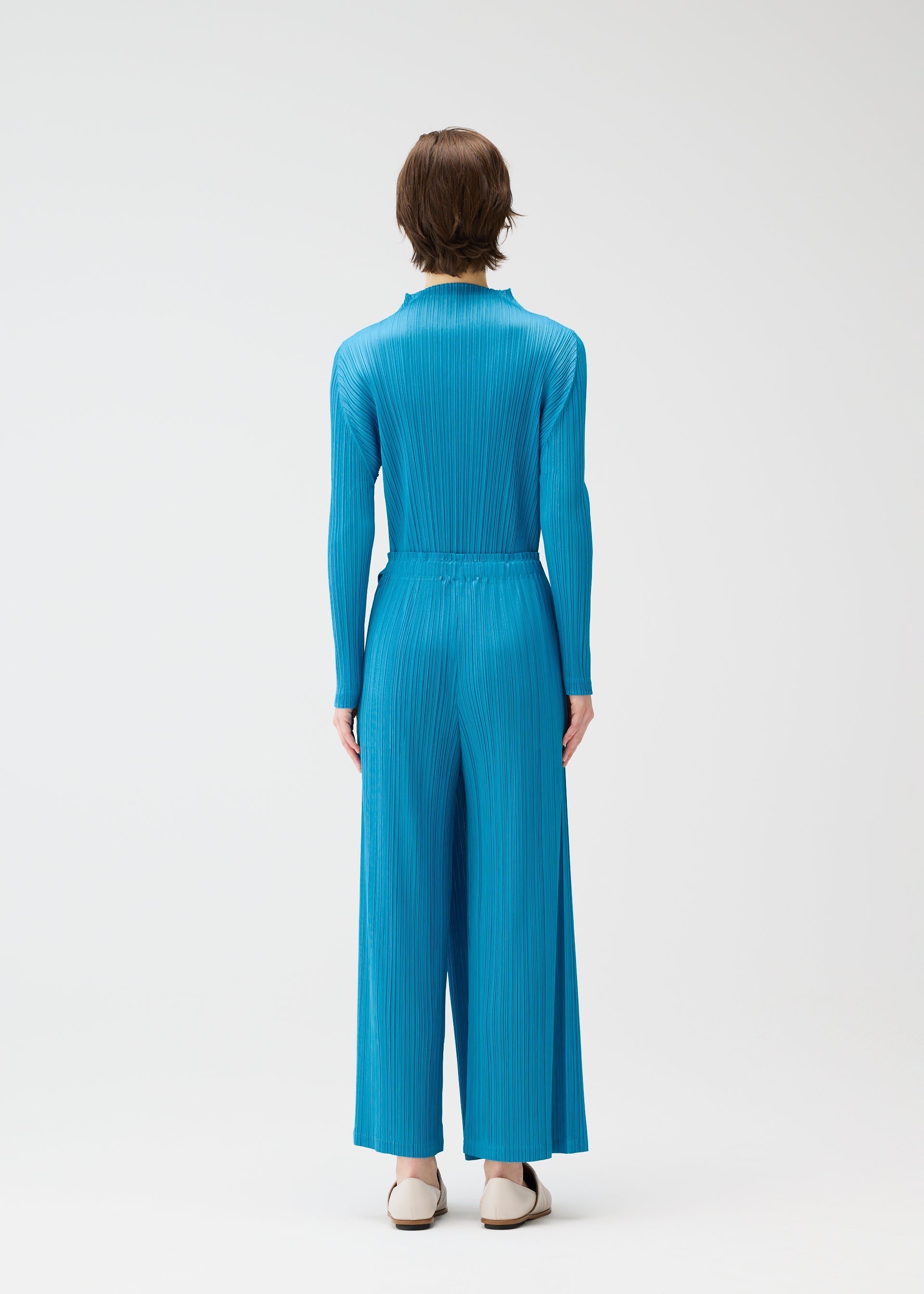 Zoomed view A person with short brown hair stands with their back to the camera, wearing the FACADE pleated jumpsuit by PLEATS PLEASE ISSEY MIYAKE and white shoes, set against a plain white background.