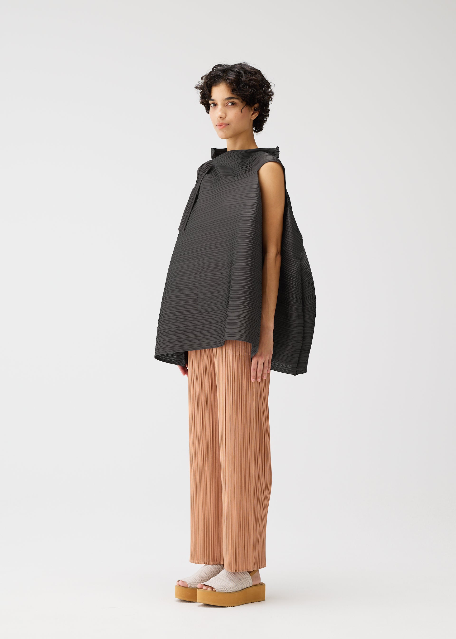 Zoomed view A person with short curly hair wears the BOUNCY BASKET top by PLEATS PLEASE ISSEY MIYAKE, pleated peach pants, and beige platform sandals with socks, standing against a plain white background.