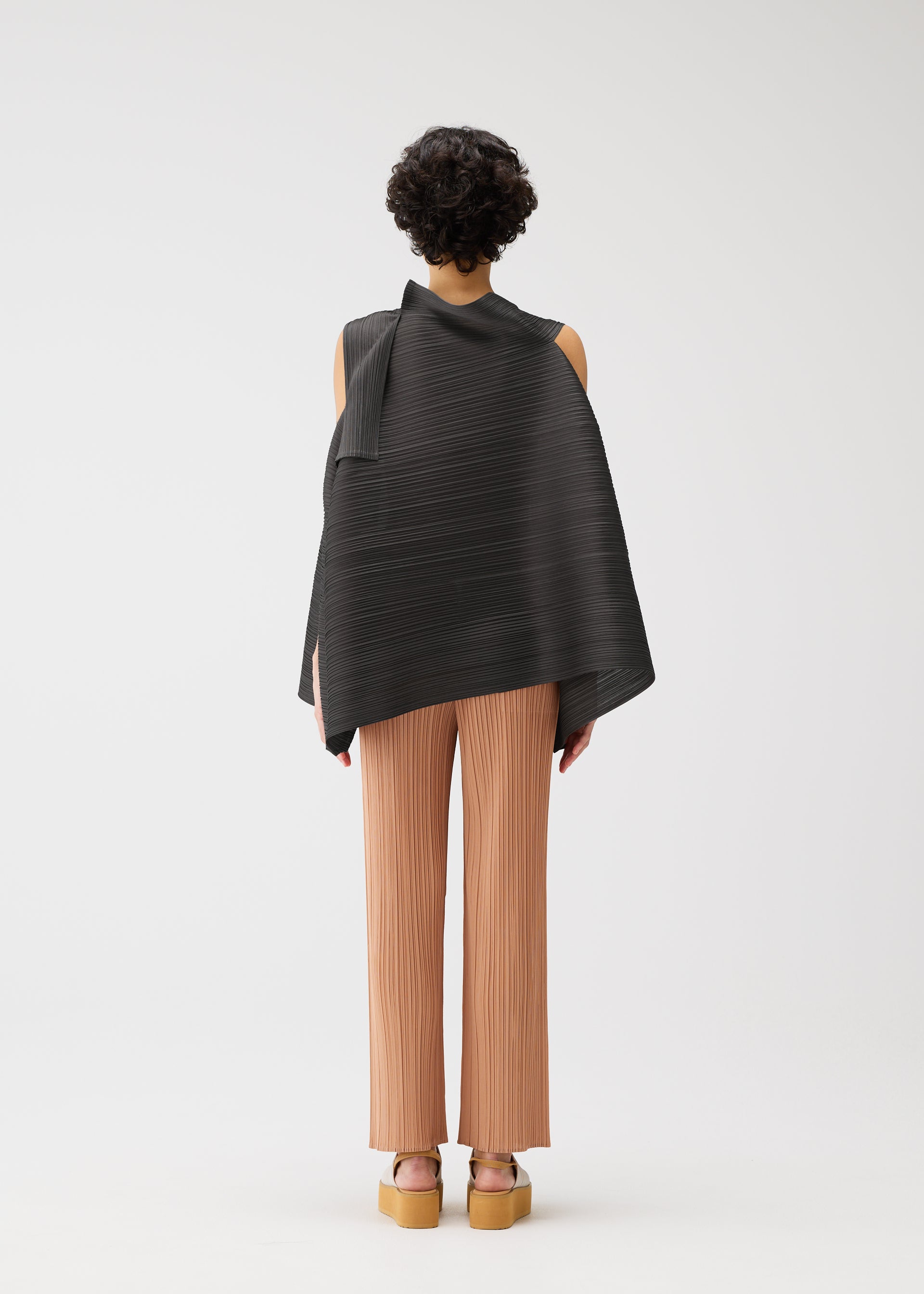 Zoomed view A person with short curly hair faces away, wearing the BOUNCY BASKET sleeveless top by PLEATS PLEASE ISSEY MIYAKE, styled with light brown pleated pants and platform sandals, set against a plain white background.
