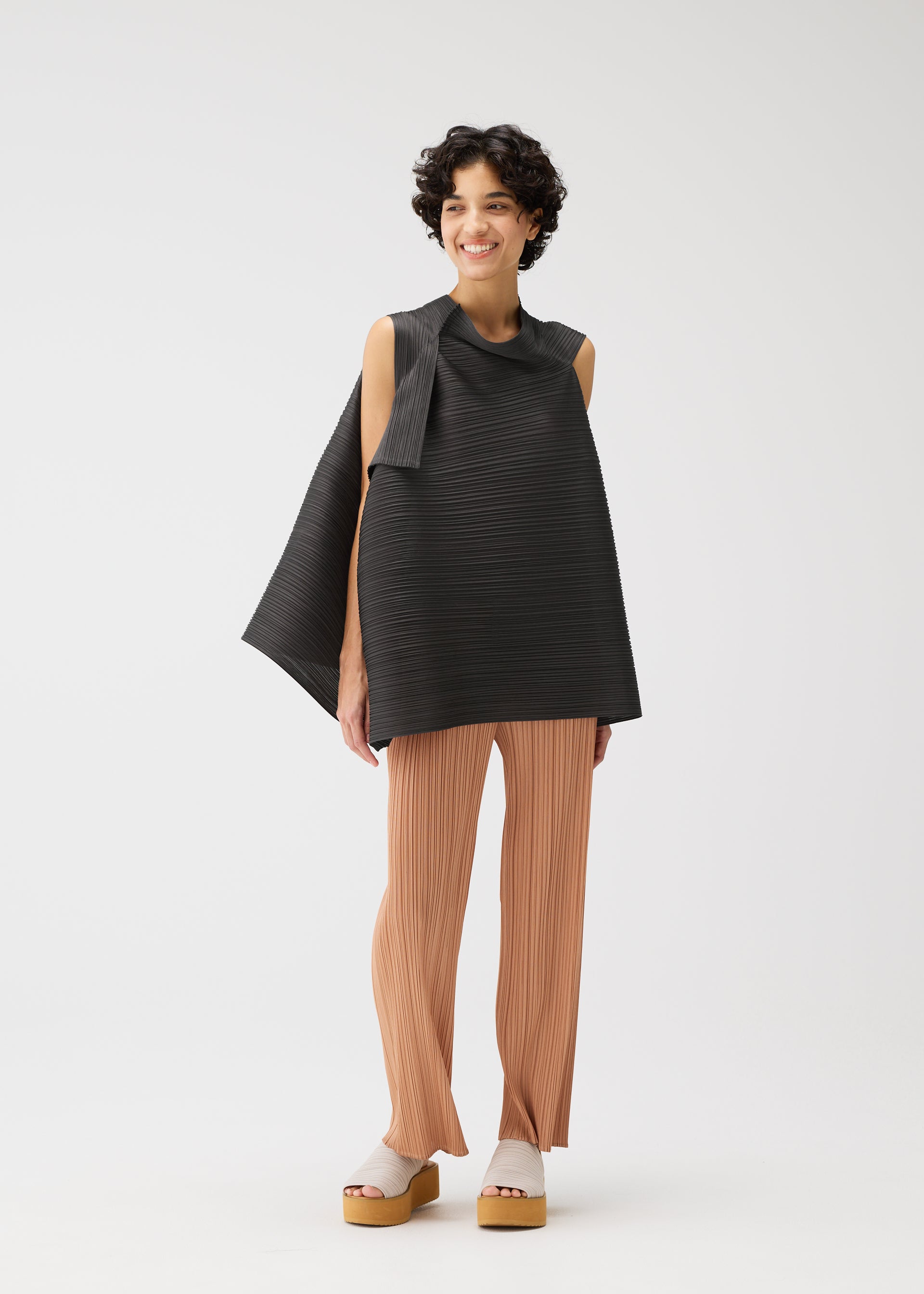 Zoomed view A person with short curly hair smiles, wearing the BOUNCY BASKET sleeveless top with unique shoulder straps and loose pleated beige pants by PLEATS PLEASE ISSEY MIYAKE, paired with light platform sandals on a white background.