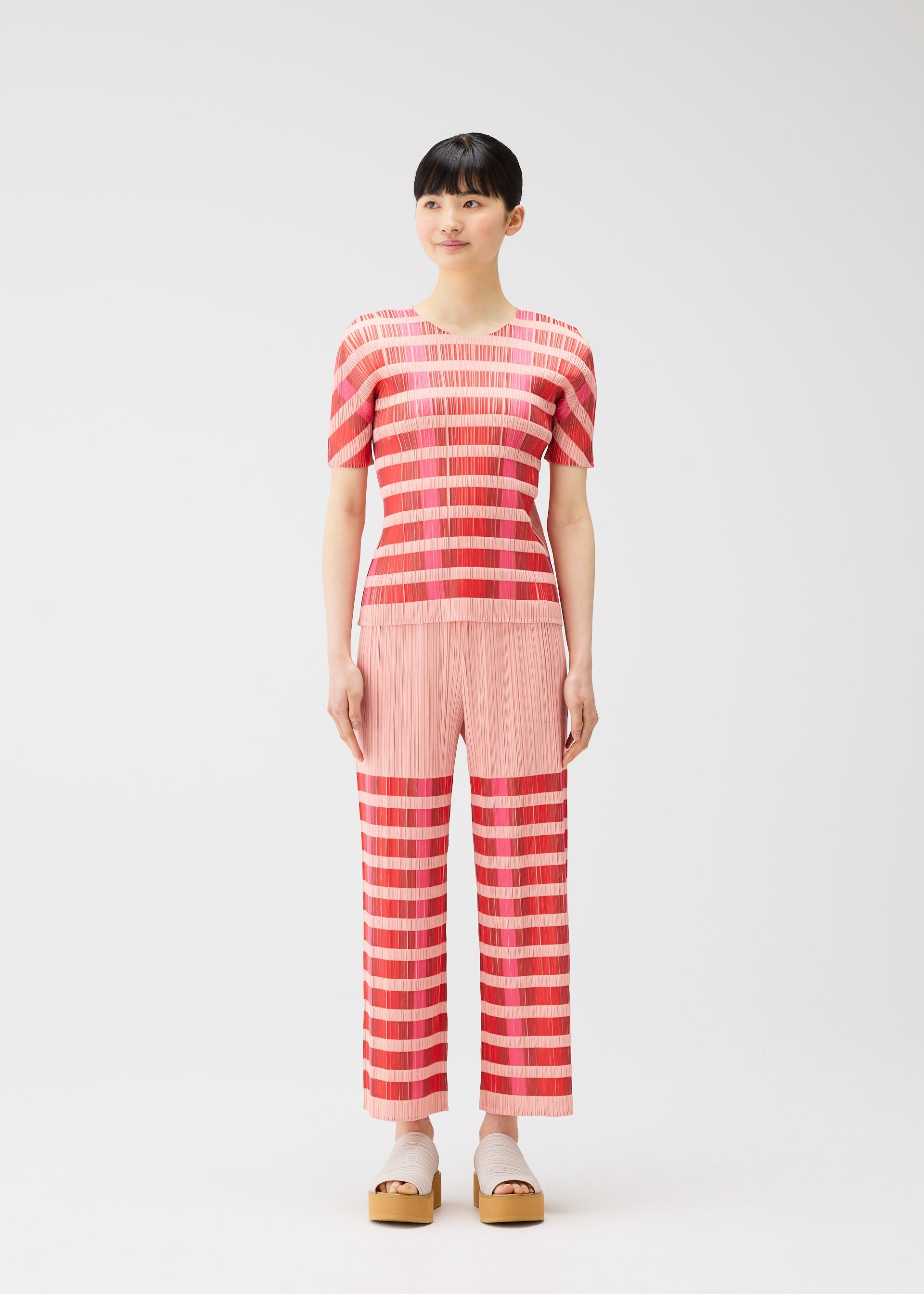 Zoomed view A person wears the PLEATS PLEASE ISSEY MIYAKE BUMPY BASKET set—short-sleeved, round-neck red and pink striped top with matching pants—styled with beige platform sandals and white socks against a white background.