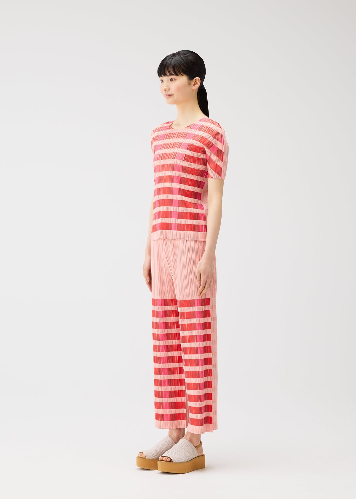 A woman with a ponytail models the BUMPY BASKET set by PLEATS PLEASE ISSEY MIYAKE: a matching pink and red round-neck, short-sleeved plaid top and wide-leg pants, paired with beige platform sandals and socks against a white background. view 6 of 8