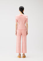A person with long black hair faces away, wearing the BUMPY BASKET pleated set by PLEATS PLEASE ISSEY MIYAKE and tan platform sandals, against a plain white background.