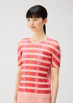 A woman with long black hair in a ponytail wears the BUMPY BASKET shirt by PLEATS PLEASE ISSEY MIYAKE, which features short sleeves and horizontal pink and red stripes. She stands against a plain light background, smiling softly.