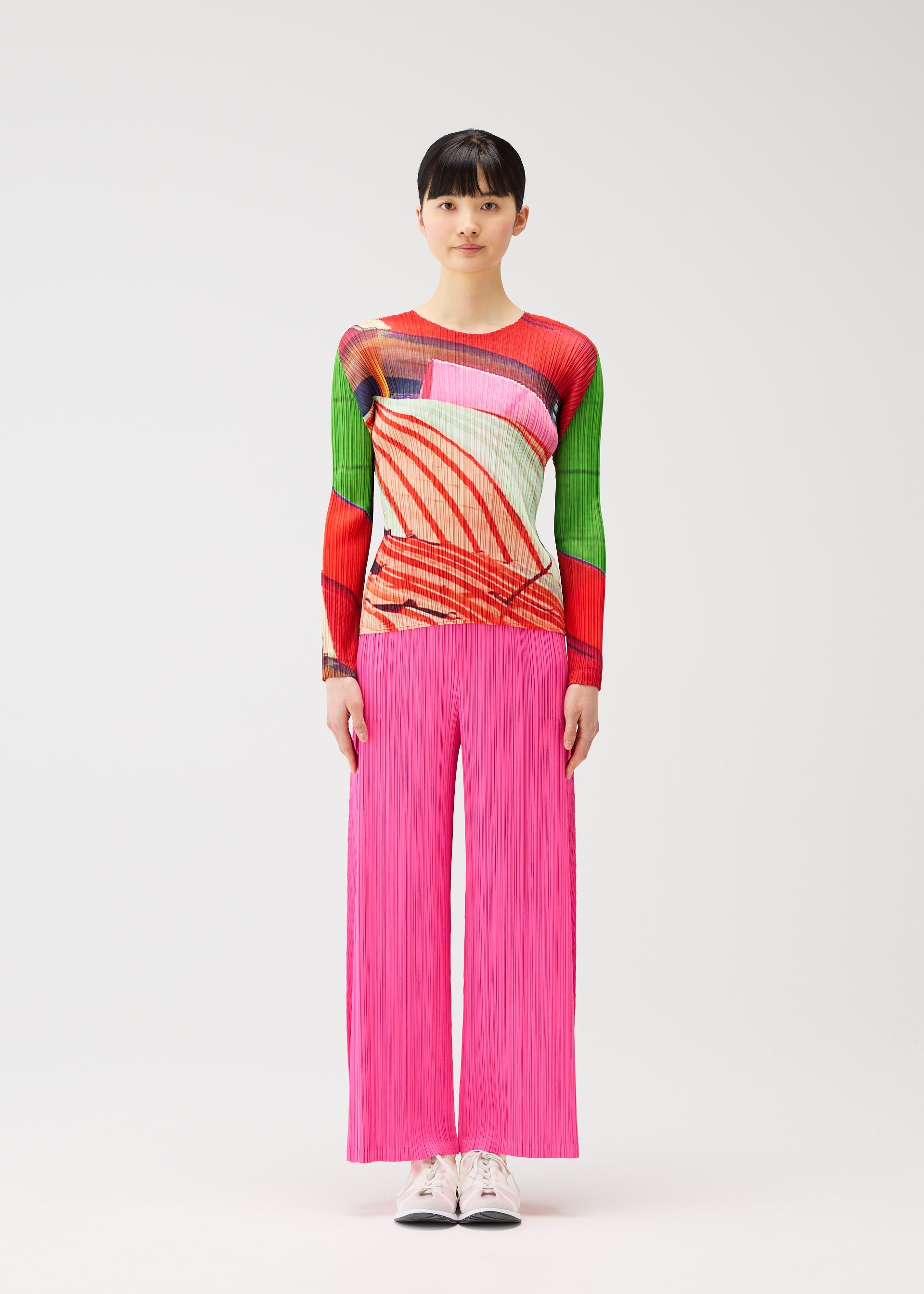Zoomed view A person with short dark hair stands against a white background wearing the CITY COLLAGE long-sleeved top with abstract patterns and pink wide-leg pleated pants by PLEATS PLEASE ISSEY MIYAKE, paired with white sneakers.