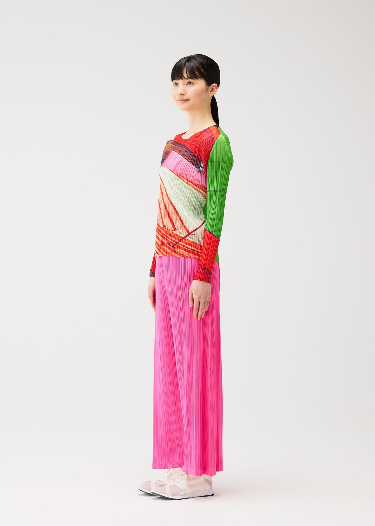 A woman stands sideways against a white background, wearing the CITY COLLAGE long-sleeved top and bright pink pleated pants by PLEATS PLEASE ISSEY MIYAKE, paired with white shoes. view 6 of 7