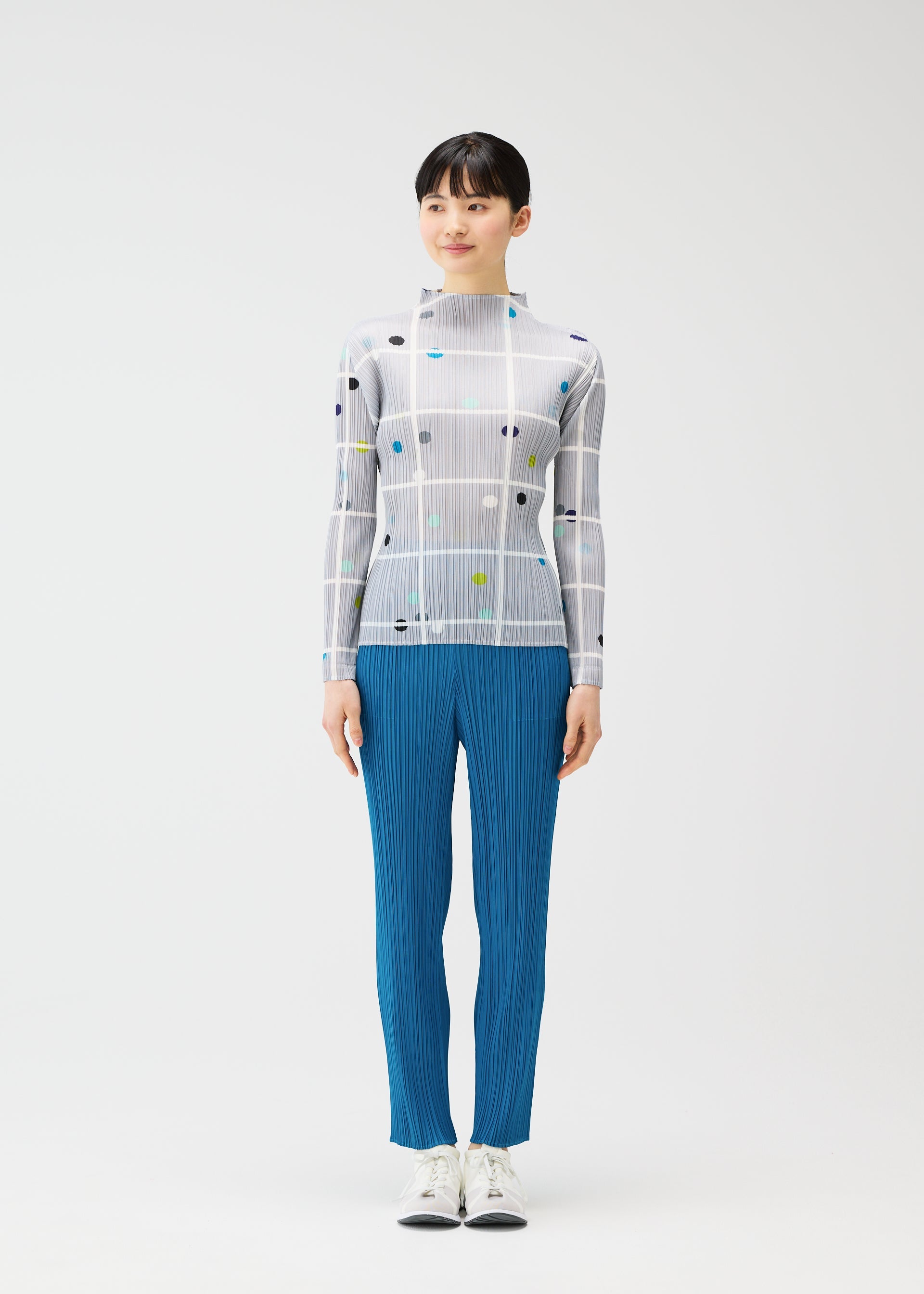 Zoomed view A person stands facing forward against a plain white background, wearing the CROSSING DOTS sheer white grid-patterned top with colored dots and blue pleated pants by PLEATS PLEASE ISSEY MIYAKE, paired with white shoes.