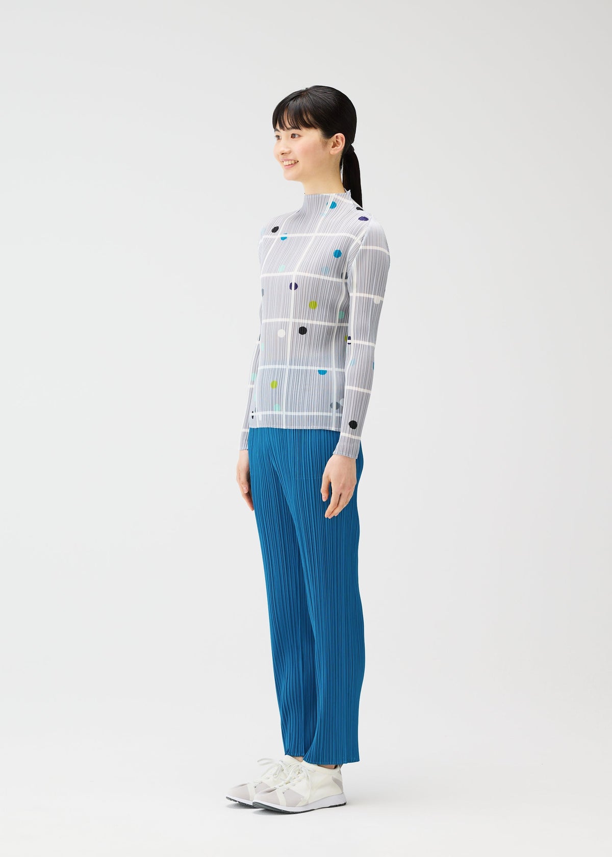 A woman stands sideways, smiling, in a fitted CROSSING DOTS long-sleeved top with colorful dots and lines by PLEATS PLEASE ISSEY MIYAKE, paired with blue pleated pants and white sneakers against a plain white background. view 5 of 6