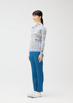 A woman stands sideways, smiling, in a fitted CROSSING DOTS long-sleeved top with colorful dots and lines by PLEATS PLEASE ISSEY MIYAKE, paired with blue pleated pants and white sneakers against a plain white background.