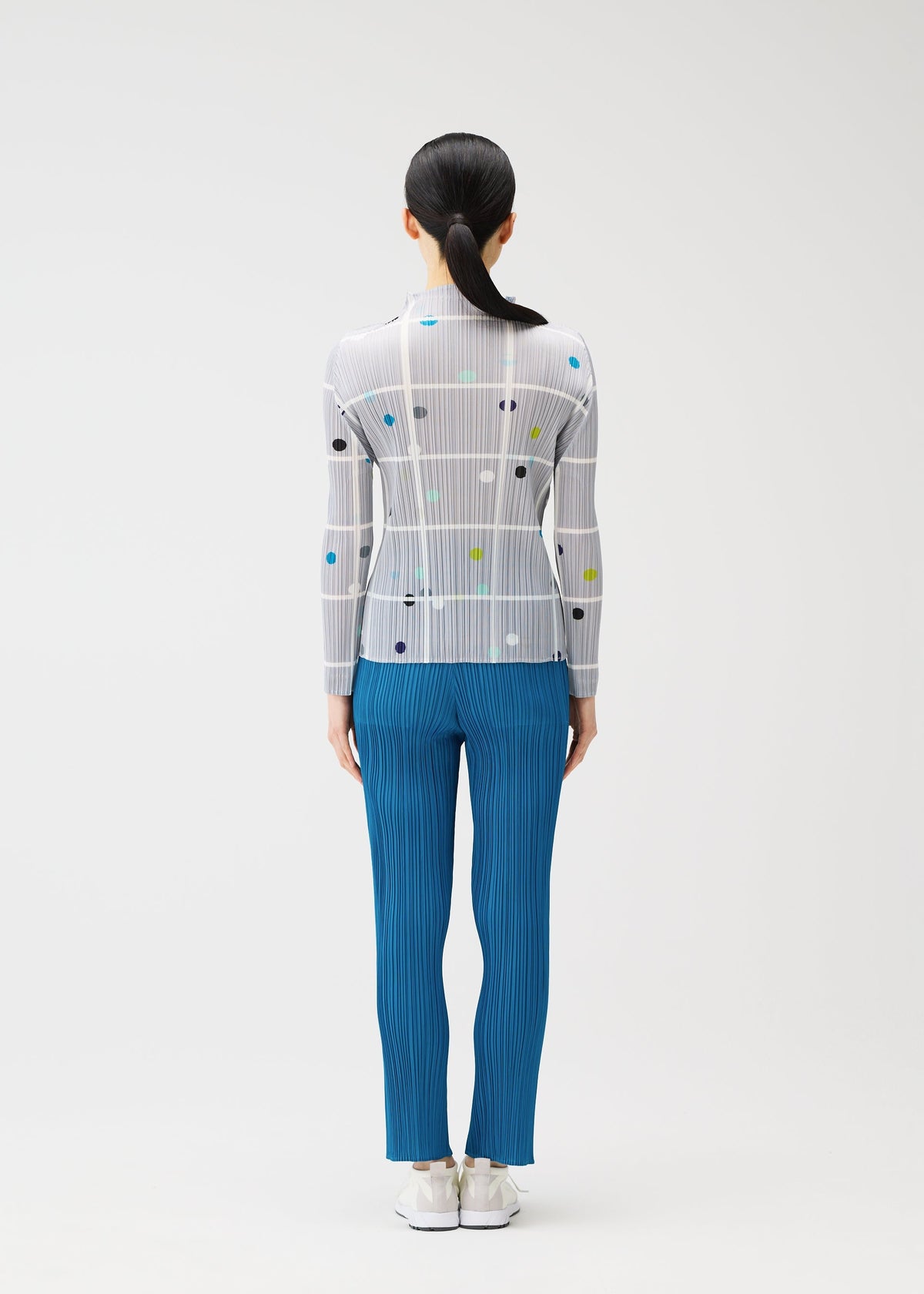 A person with long dark hair in a ponytail models the CROSSING DOTS sheer printed long-sleeved top by PLEATS PLEASE ISSEY MIYAKE, paired with blue pleated pants and white shoes, standing against a plain background. view 6 of 6
