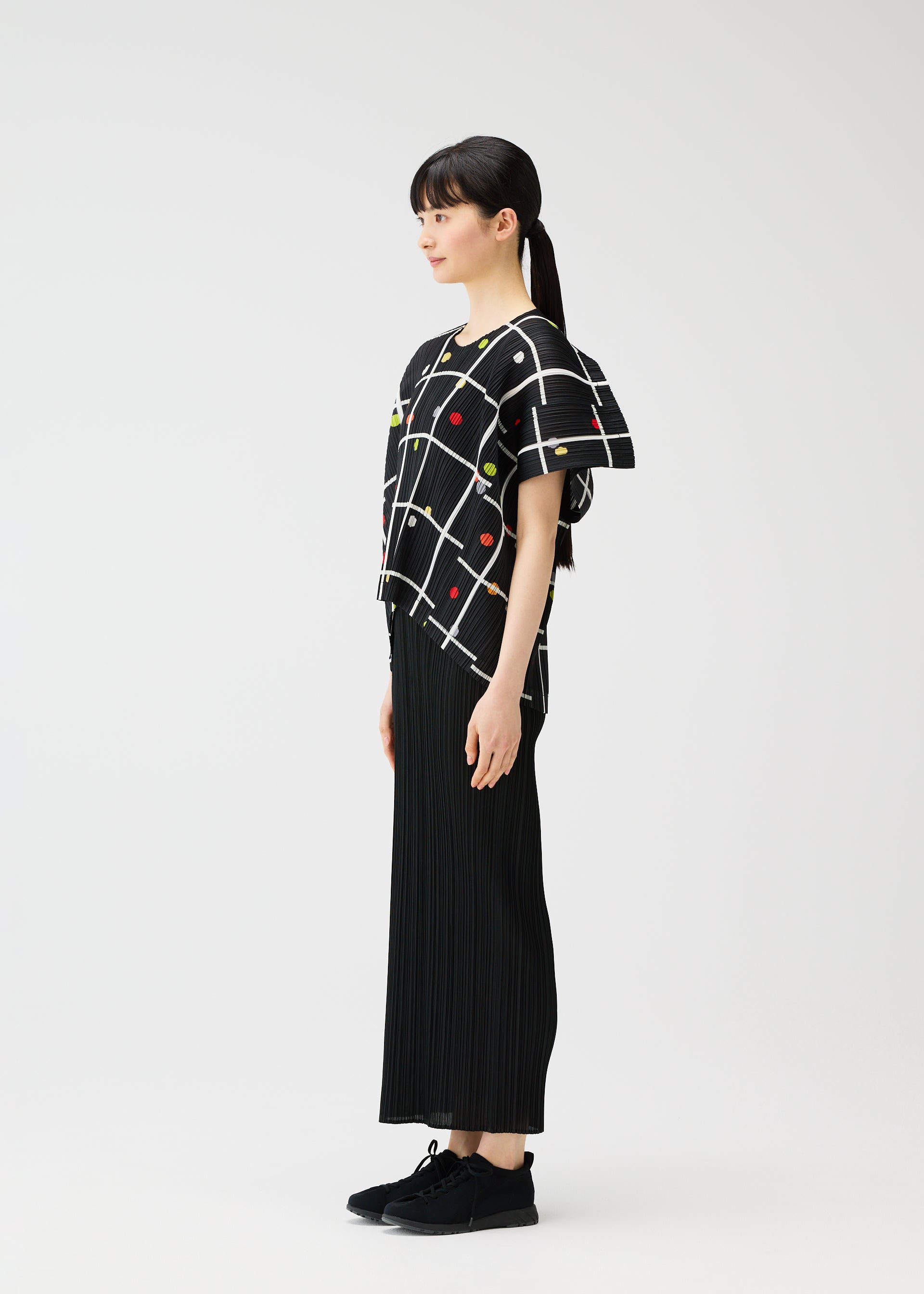 Zoomed view A woman with her hair in a ponytail stands in profile against a white background, wearing the PLEATS PLEASE ISSEY MIYAKE CROSSING DOTS round-neck top and a long black pleated skirt, paired with black sneakers.