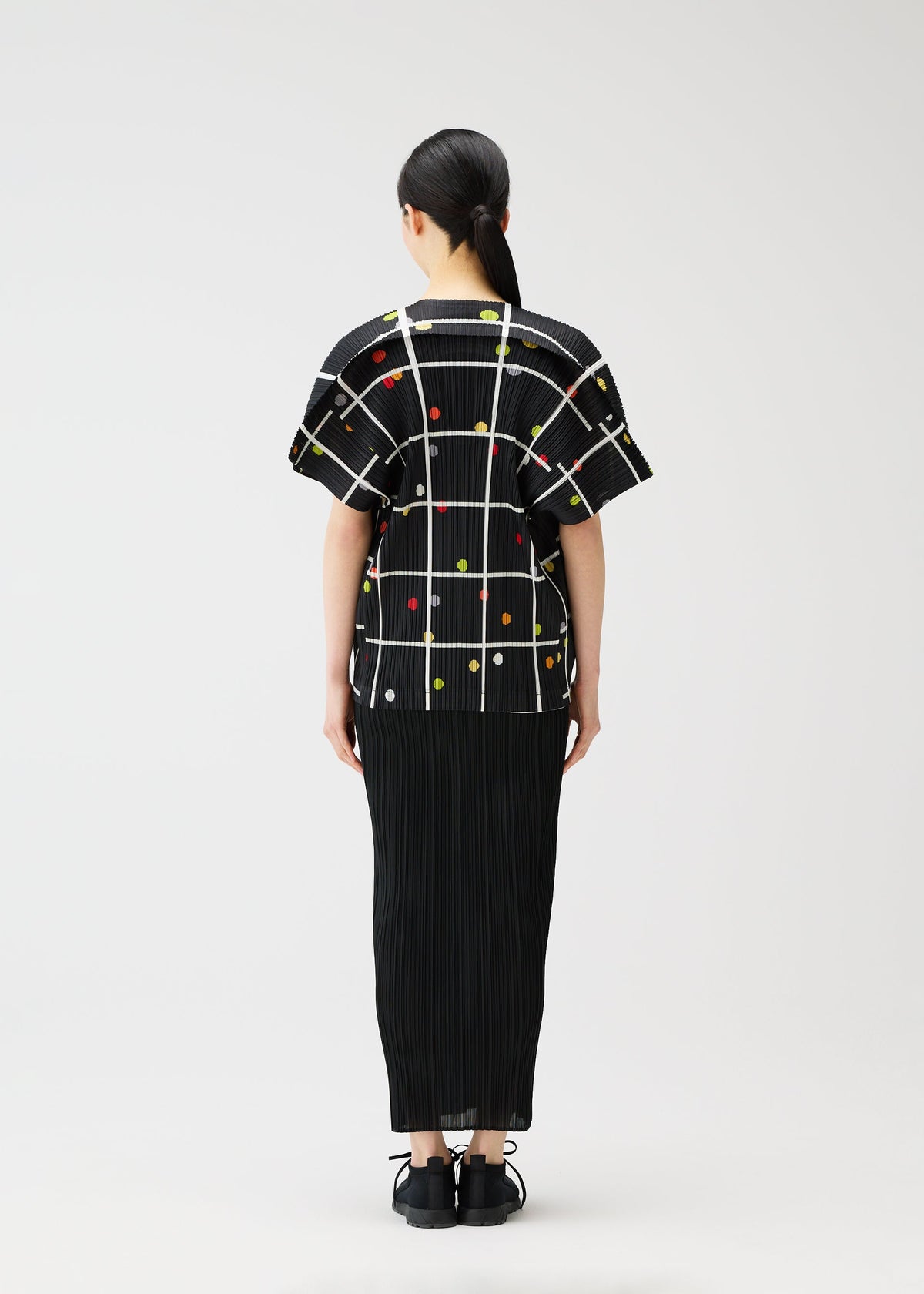 A person with dark hair in a low ponytail faces away, wearing the black textured "CROSSING DOTS" top by PLEATS PLEASE ISSEY MIYAKE with a grid and colorful dots, paired with black shoes, against a plain white background. view 8 of 10