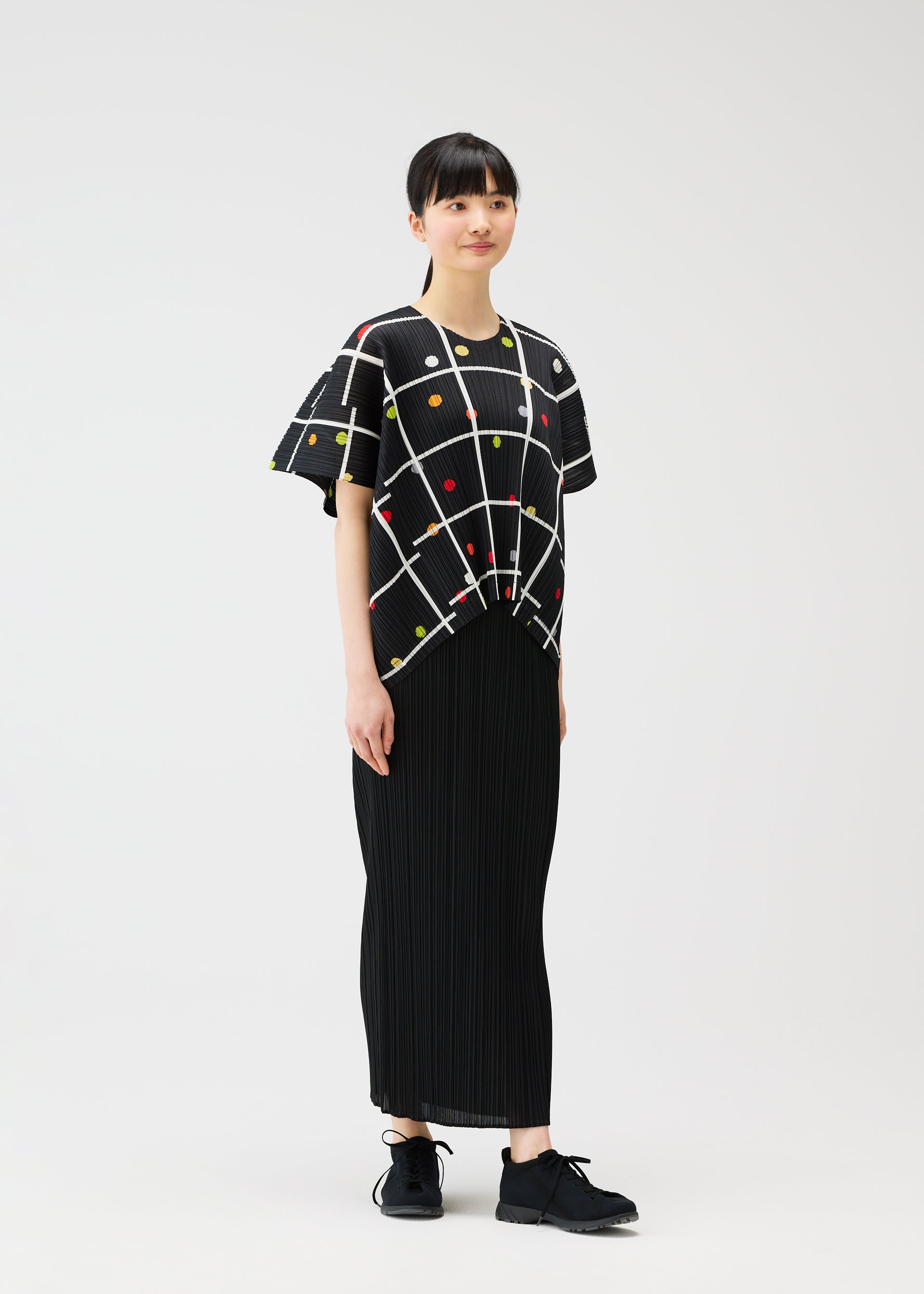Zoomed view A person wears the CROSSING DOTS top by PLEATS PLEASE ISSEY MIYAKE—a black round-neck with a geometric grid and colorful dots—paired with a long pleated skirt and black shoes, set against a white background.