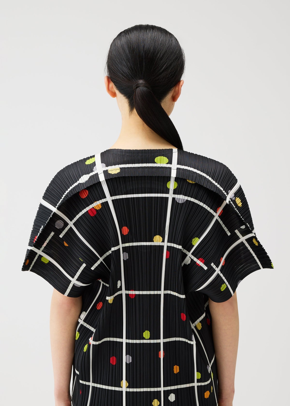 A person with straight black hair in a low ponytail is seen from behind, wearing the PLEATS PLEASE ISSEY MIYAKE CROSSING DOTS top with a black plaid and dot pattern, set against a plain light background. view 10 of 10