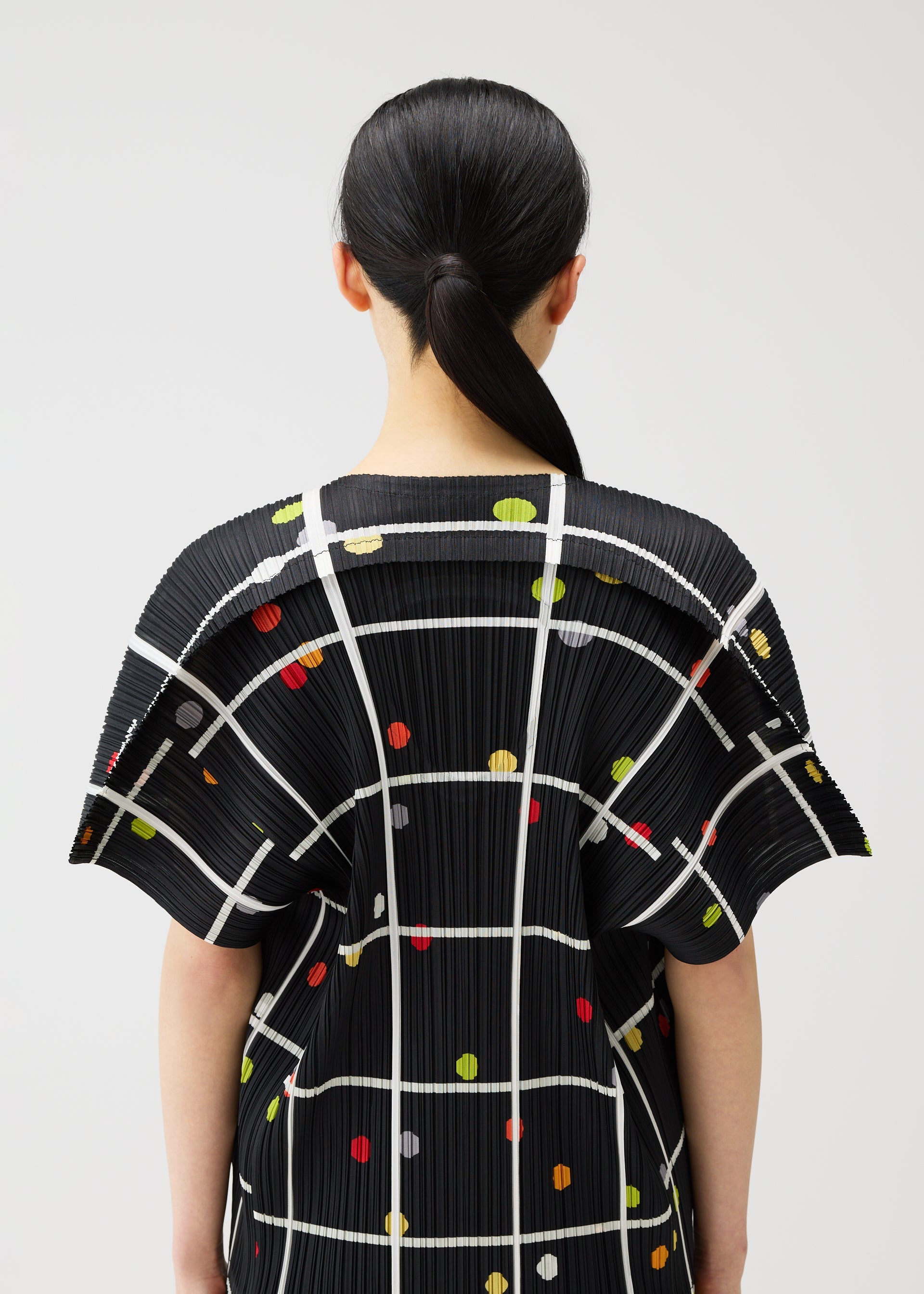 Zoomed view A person with straight black hair in a low ponytail is seen from behind, wearing the PLEATS PLEASE ISSEY MIYAKE CROSSING DOTS top with a black plaid and dot pattern, set against a plain light background.