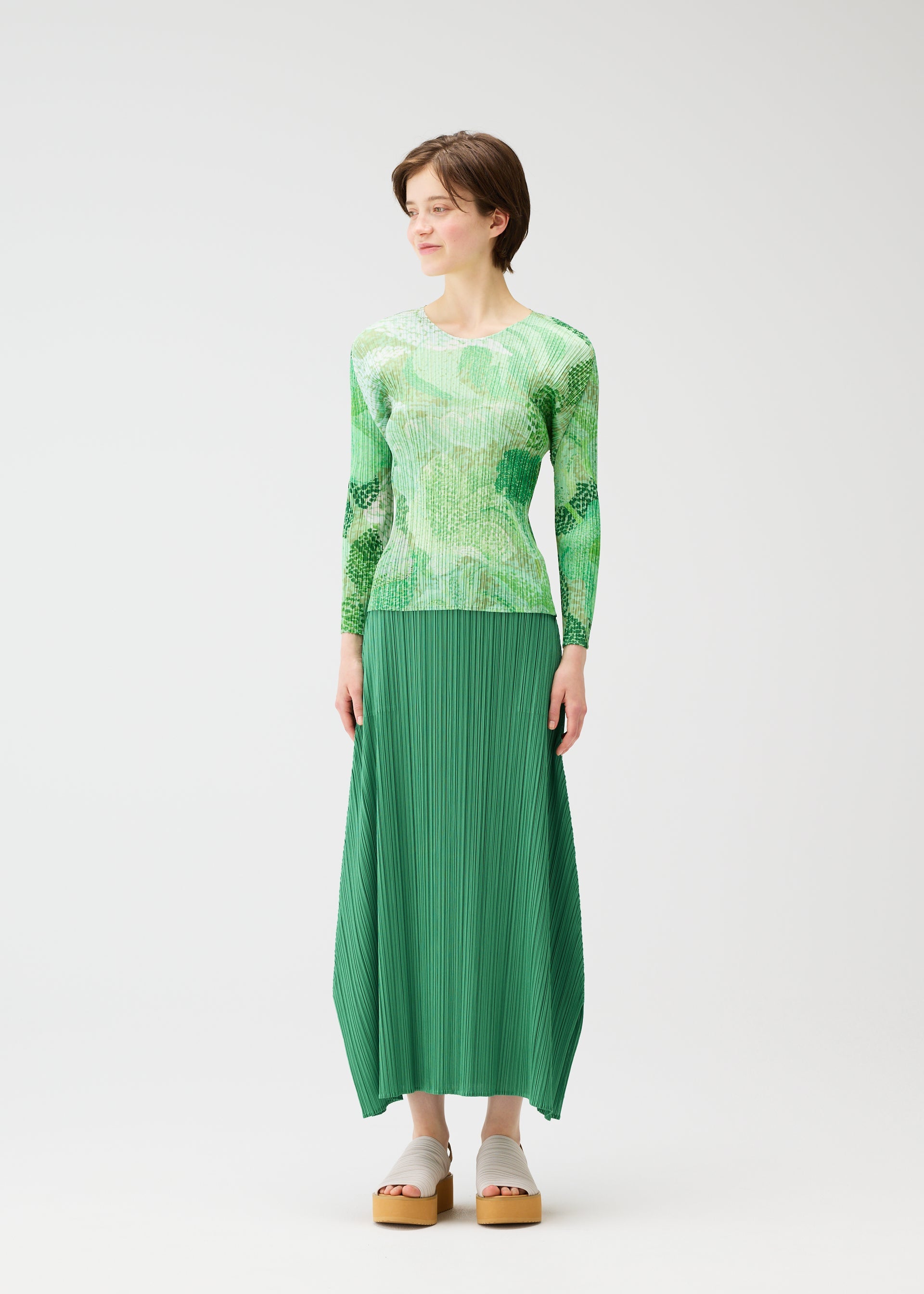Zoomed view A person wears the FOGGY FOREST top and pleated skirt by PLEATS PLEASE ISSEY MIYAKE, standing against a white background in platform sandals, looking slightly to the side with a neutral expression.
