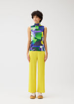 A person stands against a white background wearing the PLAYGROUND high-neck sleeveless top and bright yellow pleated pants by PLEATS PLEASE ISSEY MIYAKE, styled with beige platform sandals.