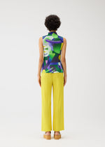 A person with short curly hair faces away, wearing the PLAYGROUND high-neck sleeveless colorful top and bright yellow pleated pants by PLEATS PLEASE ISSEY MIYAKE, paired with platform sandals against a plain light background.