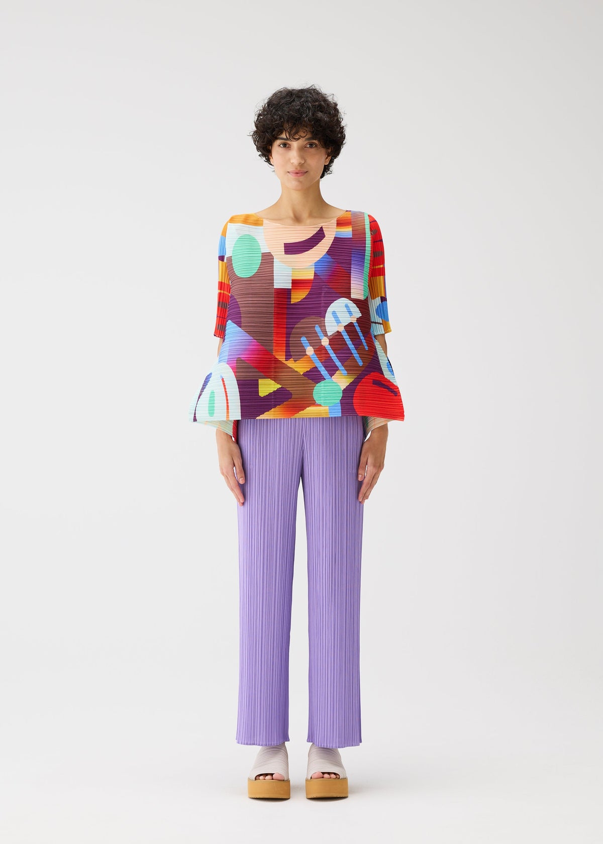 A person with short curly hair stands against a white background wearing PLEATS PLEASE ISSEY MIYAKE's PLAYGROUND geometric-print round-neck top with flared sleeves, lilac pleated pants, and platform sandals. view 6 of 9