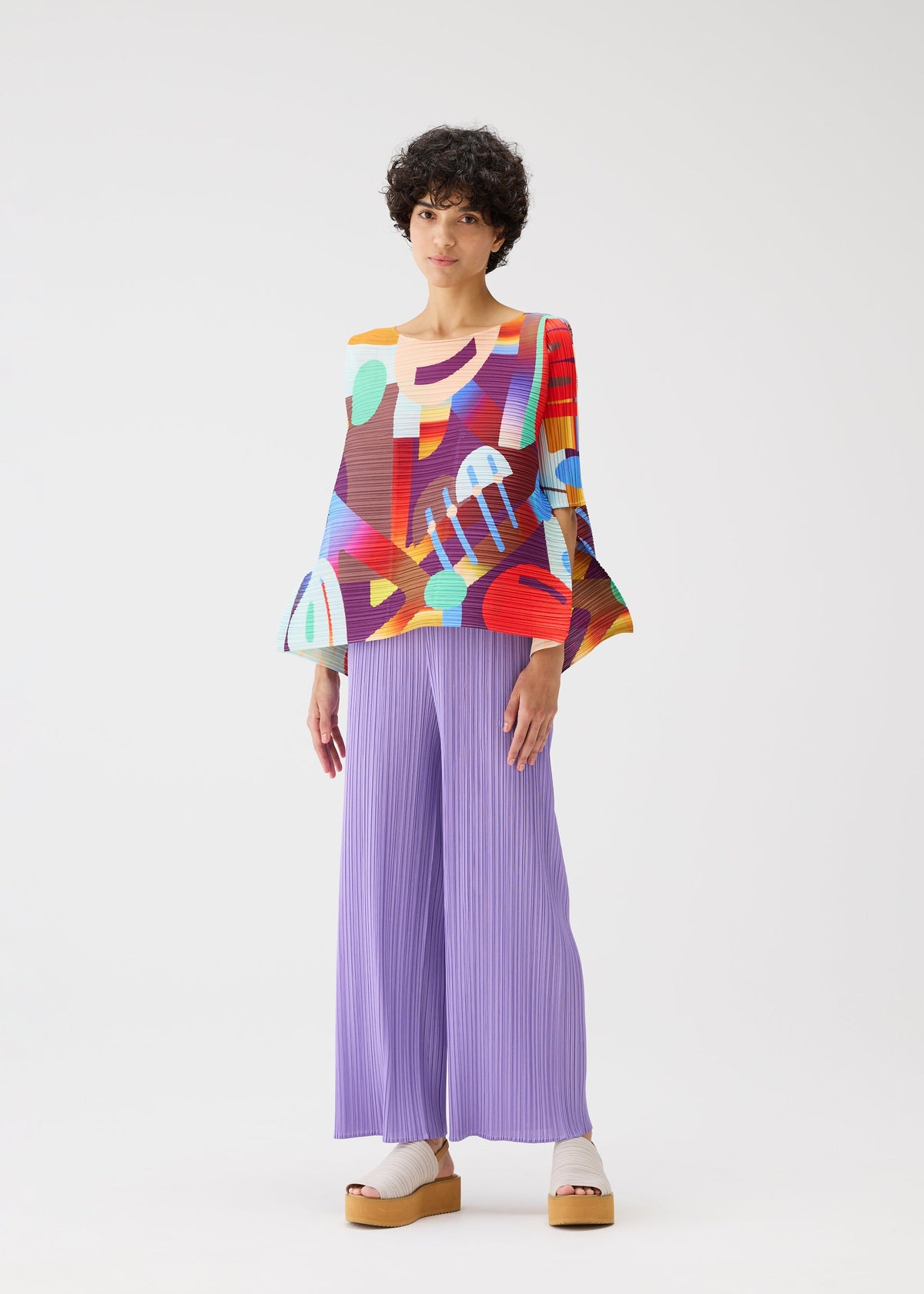 A person stands against a white background, wearing the colorful PLAYGROUND top with wide sleeves and geometric patterns from PLEATS PLEASE ISSEY MIYAKE, paired with loose pleated lavender pants and beige platform sandals. view 9 of 9