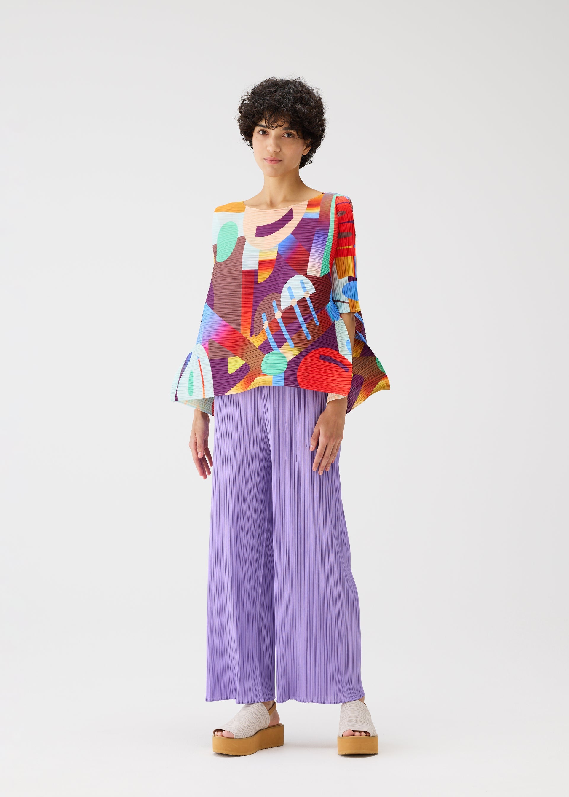 Zoomed view A person stands against a white background, wearing the colorful PLAYGROUND top with wide sleeves and geometric patterns from PLEATS PLEASE ISSEY MIYAKE, paired with loose pleated lavender pants and beige platform sandals.