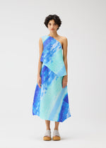 A person models the SEA BUBBLE midi dress by PLEATS PLEASE ISSEY MIYAKE—an abstract-patterned, sleeveless, blue and turquoise one-shoulder dress with diagonal pleats—facing forward against a plain white background in beige platform sandals.