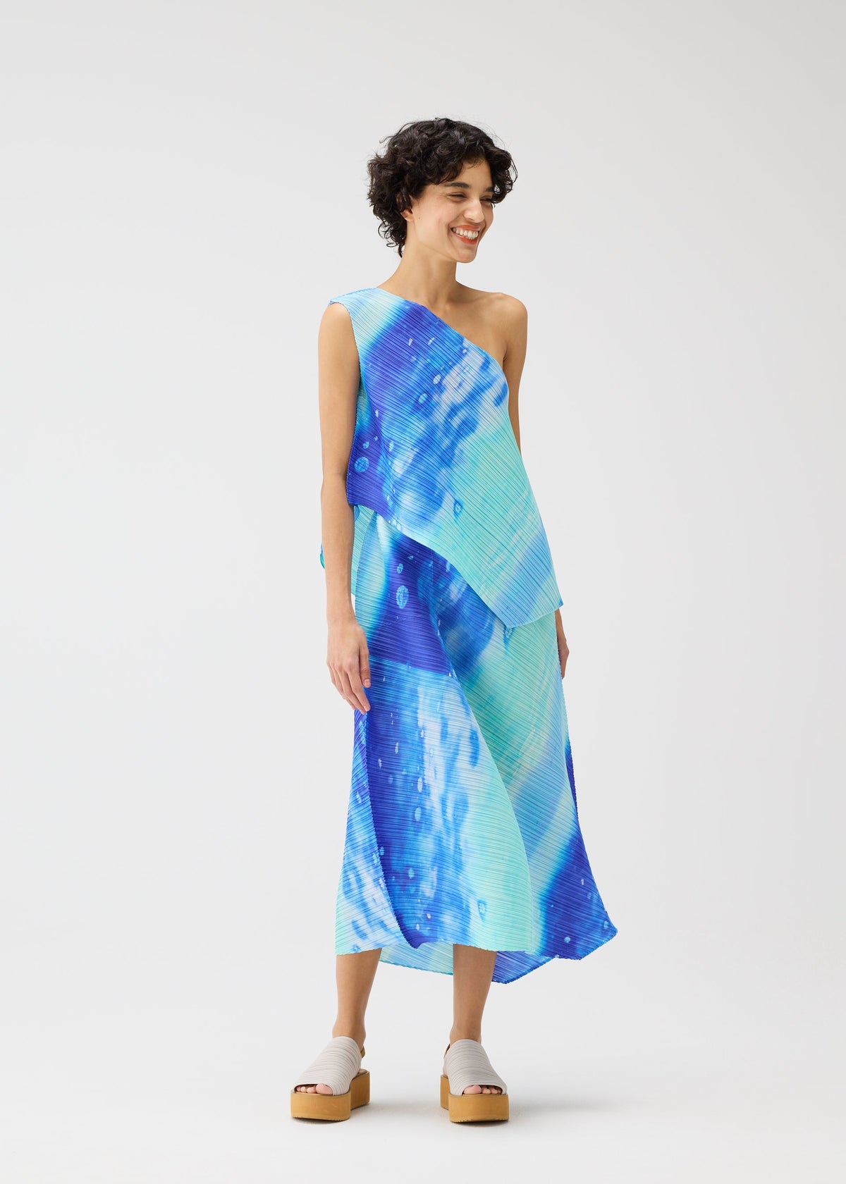 A person smiles in the SEA BUBBLE dress by PLEATS PLEASE ISSEY MIYAKE, featuring blue and teal one-shoulder asymmetry, diagonal pleats, and layered design. They have short curly hair and light platform sandals against a white background. view 7 of 7