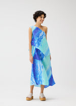 A person smiles in the SEA BUBBLE dress by PLEATS PLEASE ISSEY MIYAKE, featuring blue and teal one-shoulder asymmetry, diagonal pleats, and layered design. They have short curly hair and light platform sandals against a white background.