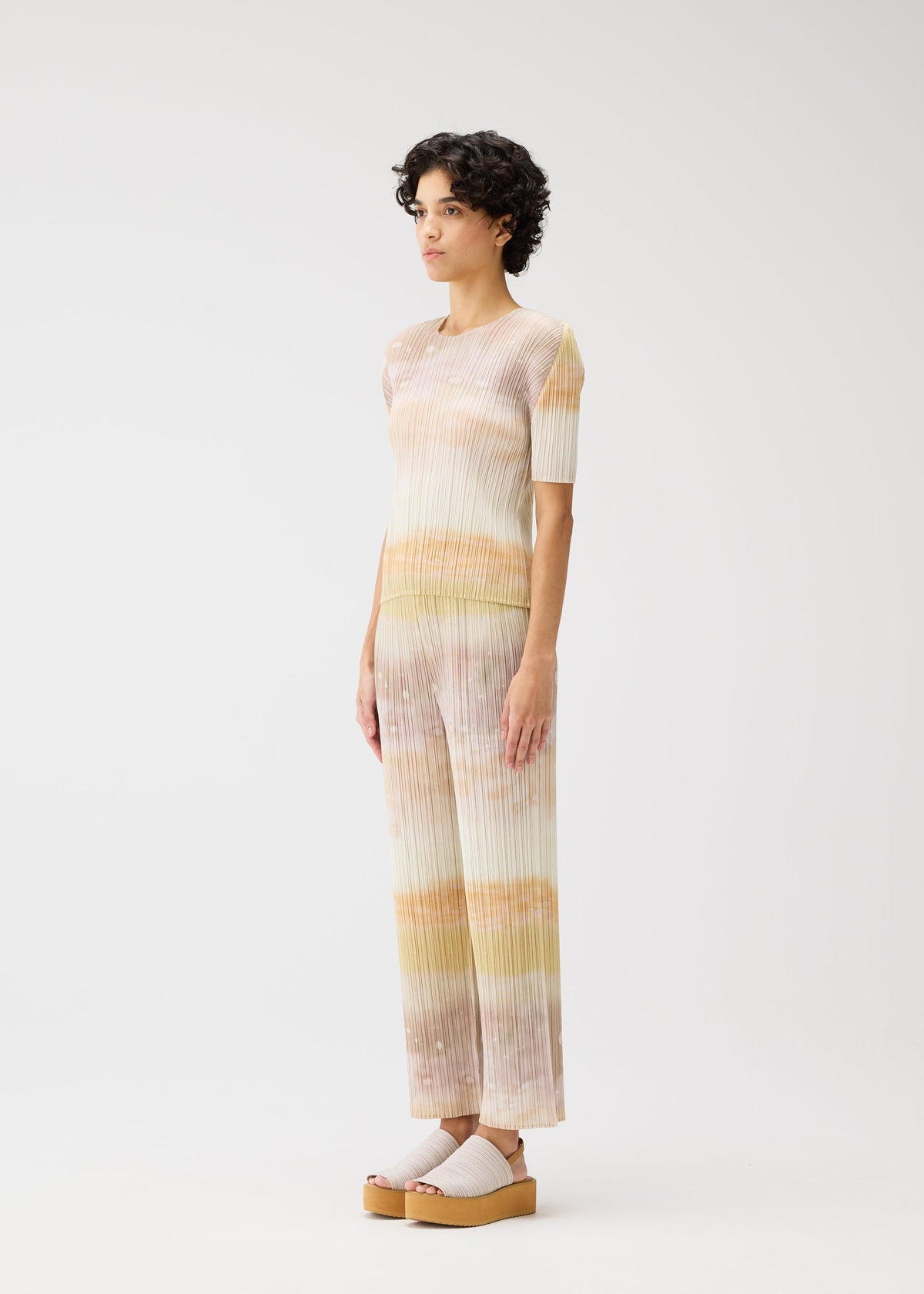 A person with short curly hair wears the SEA BUBBLE set by PLEATS PLEASE ISSEY MIYAKE—pastel-striped, short-sleeved top and matching wide-leg pants with a subtle beach pattern—plus light platform sandals, posed against a plain white background. view 5 of 6