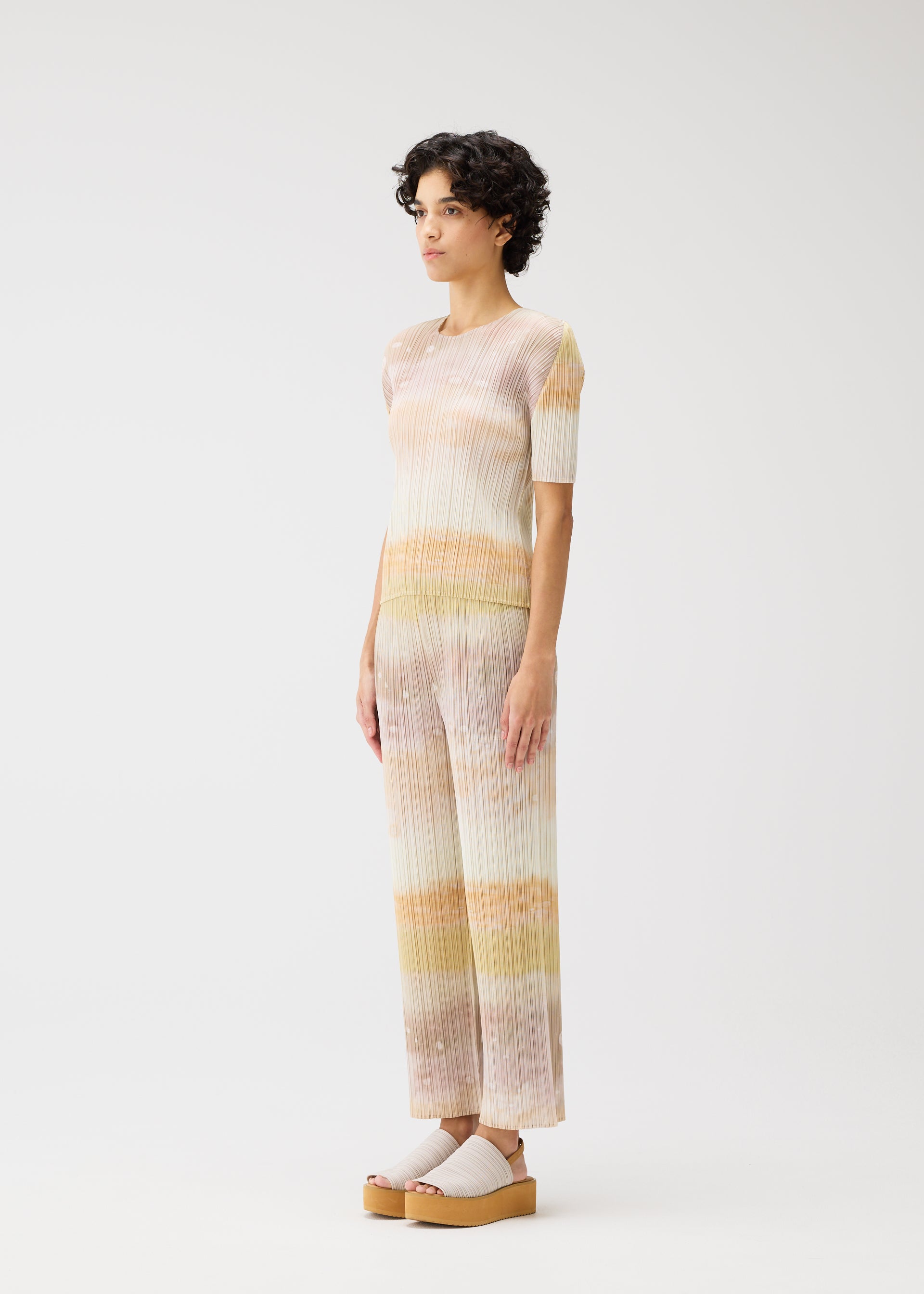 Zoomed view A person with short curly hair wears the SEA BUBBLE set by PLEATS PLEASE ISSEY MIYAKE—pastel-striped, short-sleeved top and matching wide-leg pants with a subtle beach pattern—plus light platform sandals, posed against a plain white background.