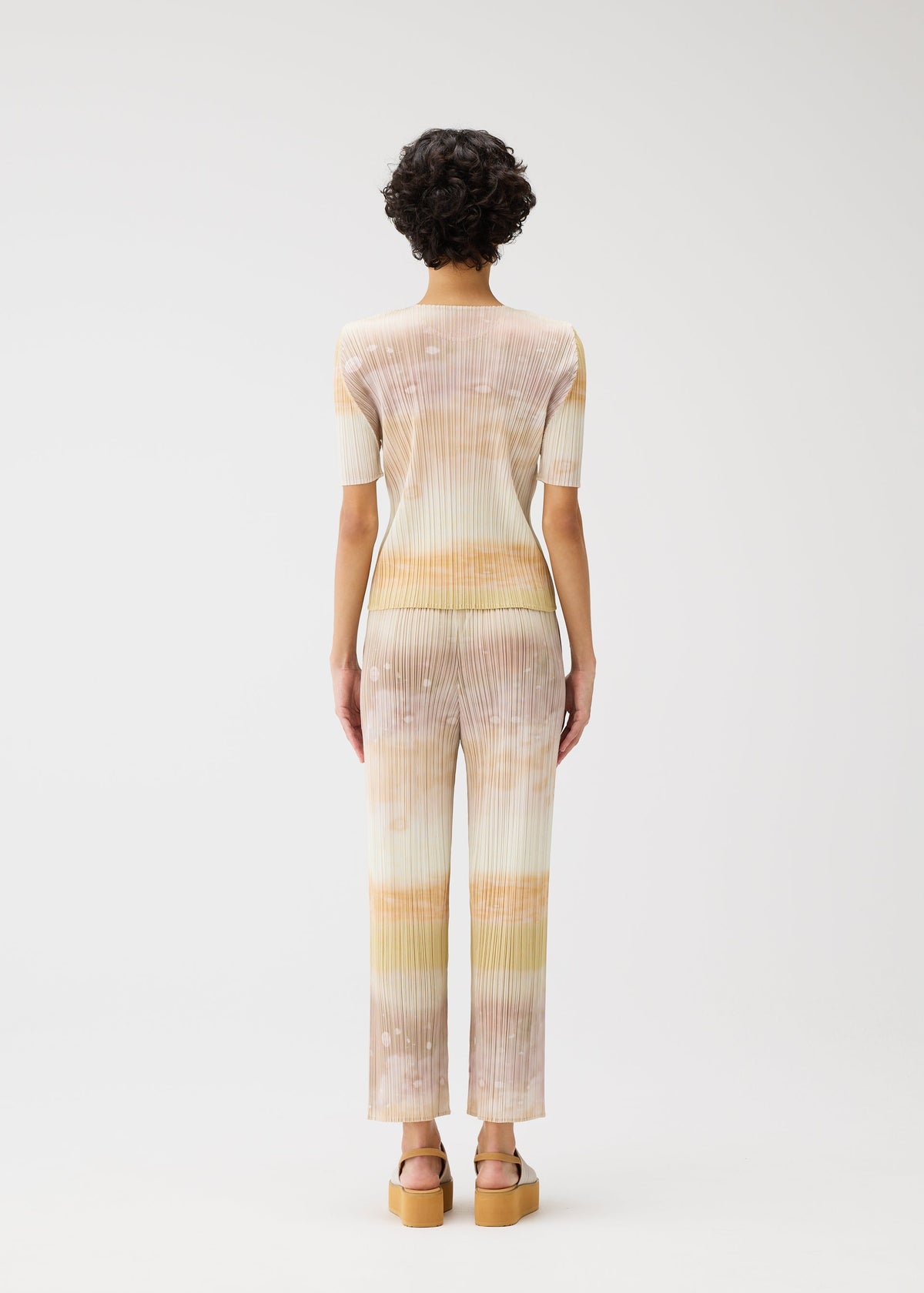 A person with short curly hair faces away, wearing the SEA BUBBLE set by PLEATS PLEASE ISSEY MIYAKE—pastel striped round-neck top with short sleeves and matching flared pants—styled with tan platform sandals against a white background. view 6 of 6