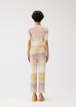 A person with short curly hair faces away, wearing the SEA BUBBLE set by PLEATS PLEASE ISSEY MIYAKE—pastel striped round-neck top with short sleeves and matching flared pants—styled with tan platform sandals against a white background.