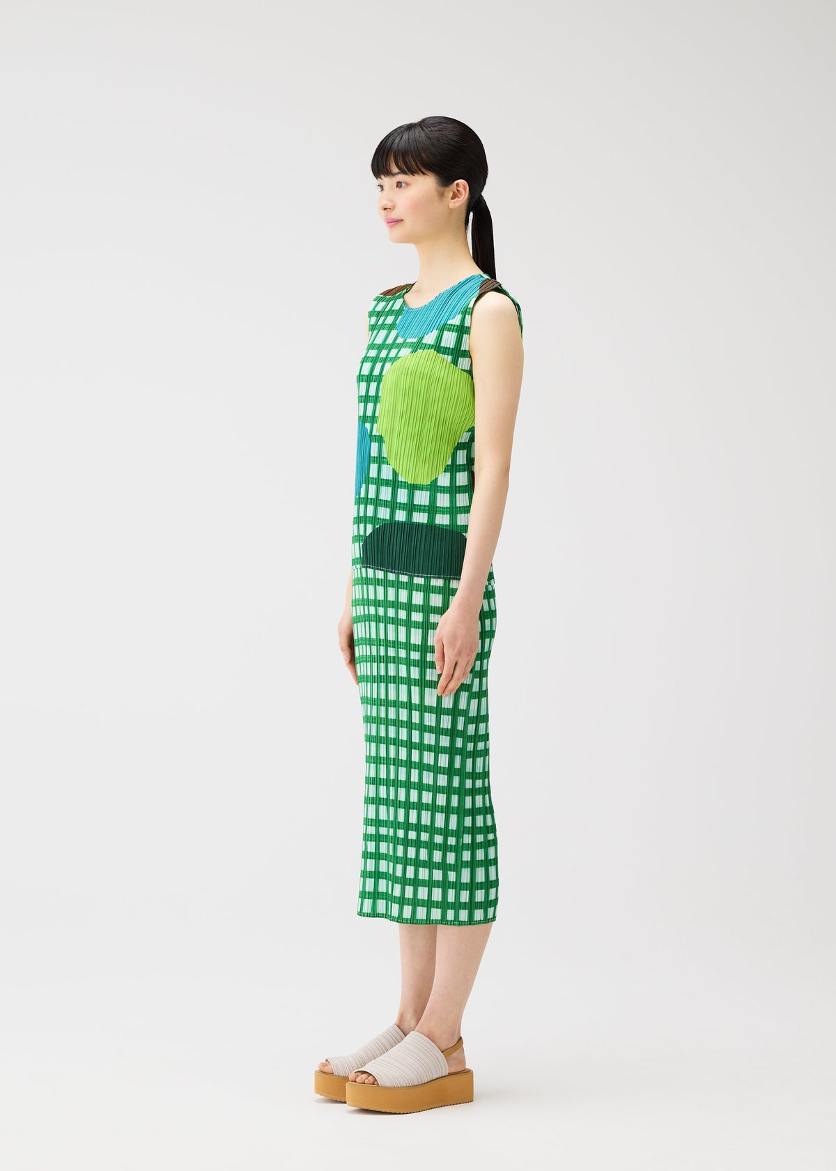 A woman with straight black hair in a ponytail stands sideways, holding the MARKET basket from PLEATS PLEASE ISSEY MIYAKE. She wears a sleeveless green and white checkered dress with large green circles and beige platform sandals against a white background. view 5 of 6