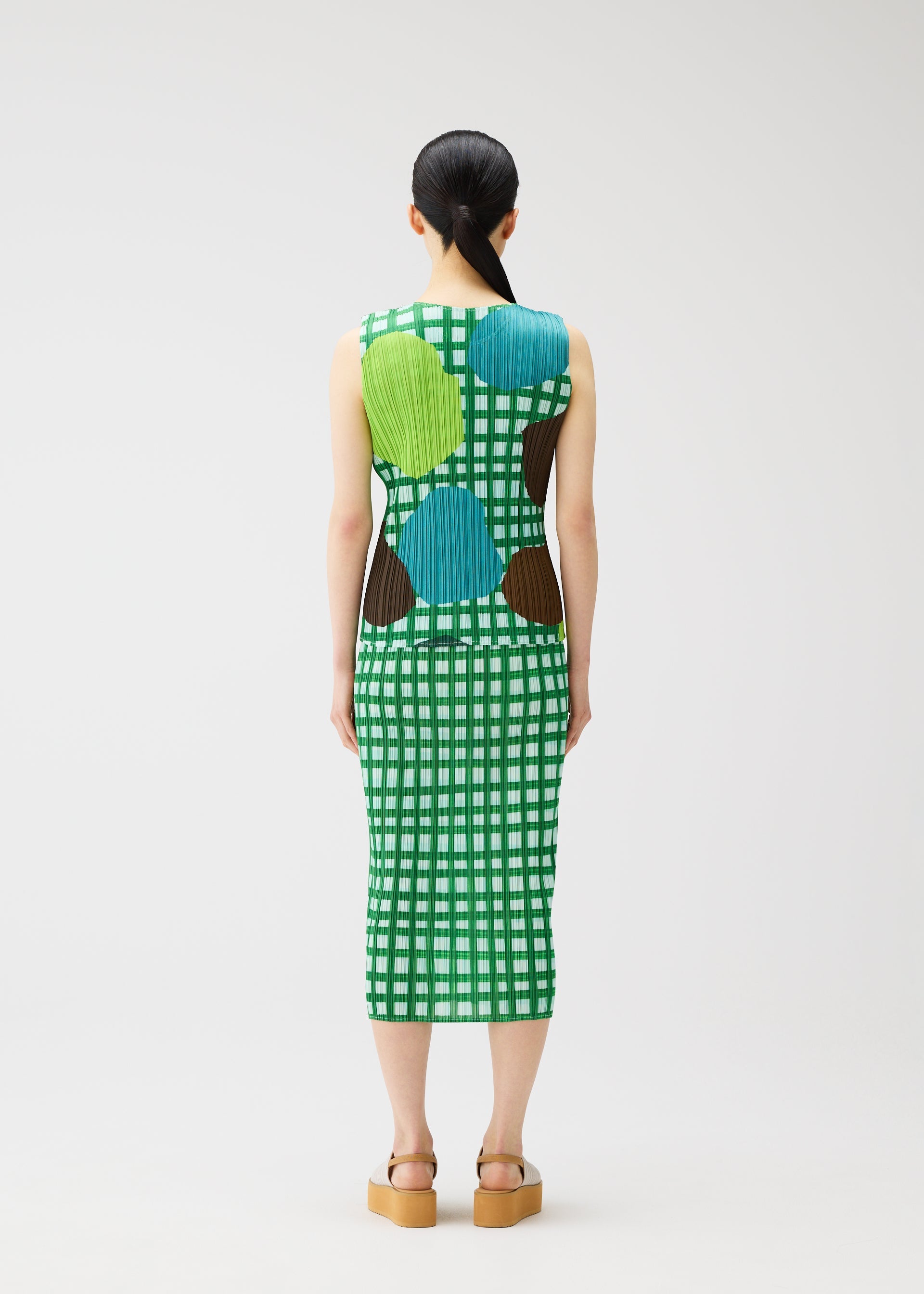 Zoomed view A person with long dark hair in a ponytail models the MARKET dress by PLEATS PLEASE ISSEY MIYAKE—a sleeveless, calf-length green and white checkered dress with blue, green, and brown circular patterns—paired with tan platform sandals.