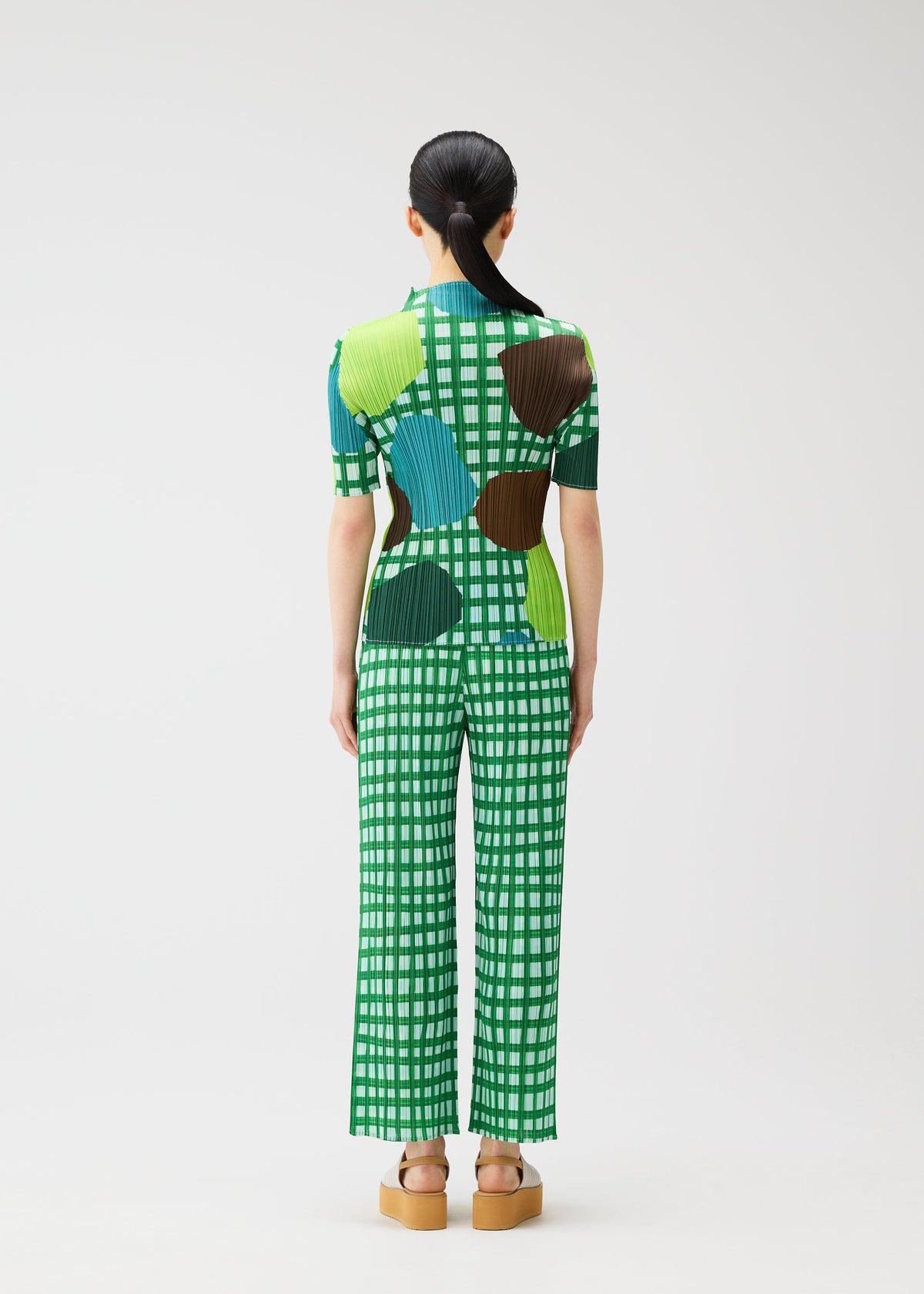 A person with long black hair in a low ponytail stands facing away, wearing the MARKET outfit by PLEATS PLEASE ISSEY MIYAKE featuring a green and white checkered playful vegetable fruit pattern and tan platform sandals against a white background. view 6 of 6