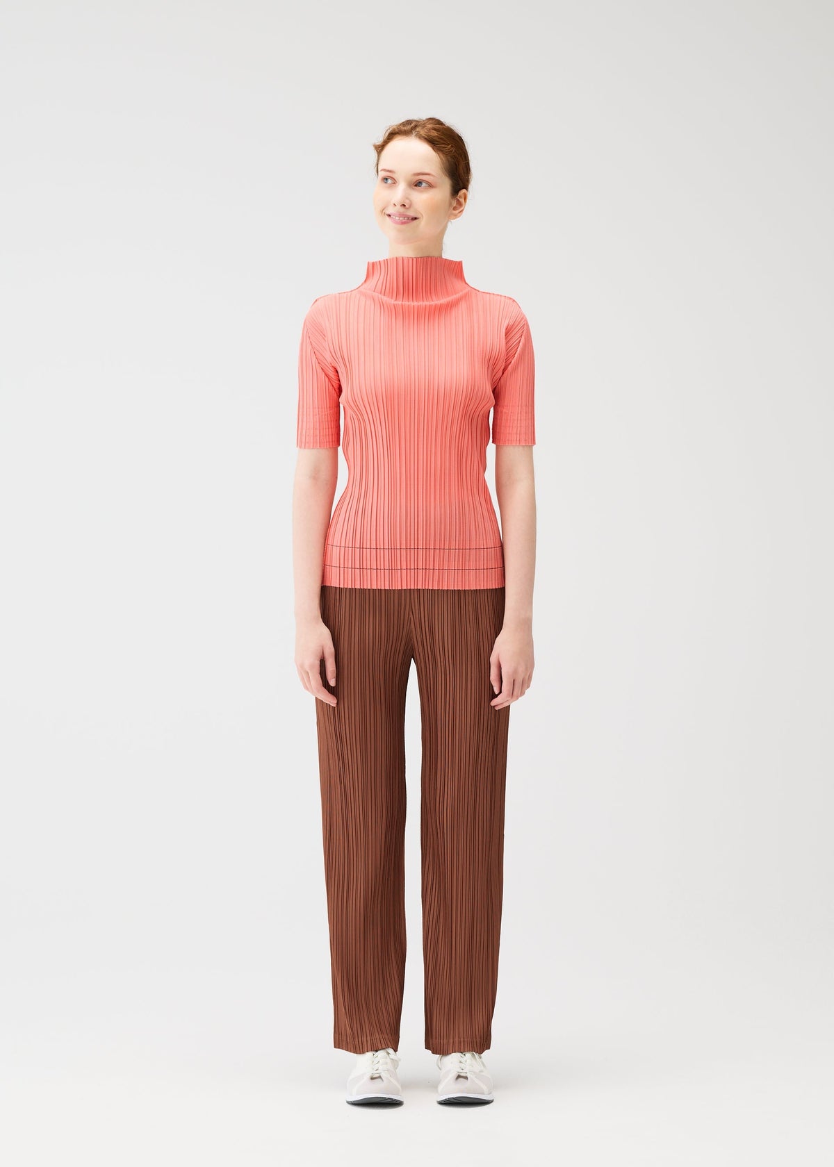 A woman stands against a light background, wearing the SOFT PLEATS pants by PLEATS PLEASE ISSEY MIYAKE with a pink ribbed high-neck top and white sneakers—an effortlessly chic spring look. She glances sideways, smiling softly. view 5 of 7