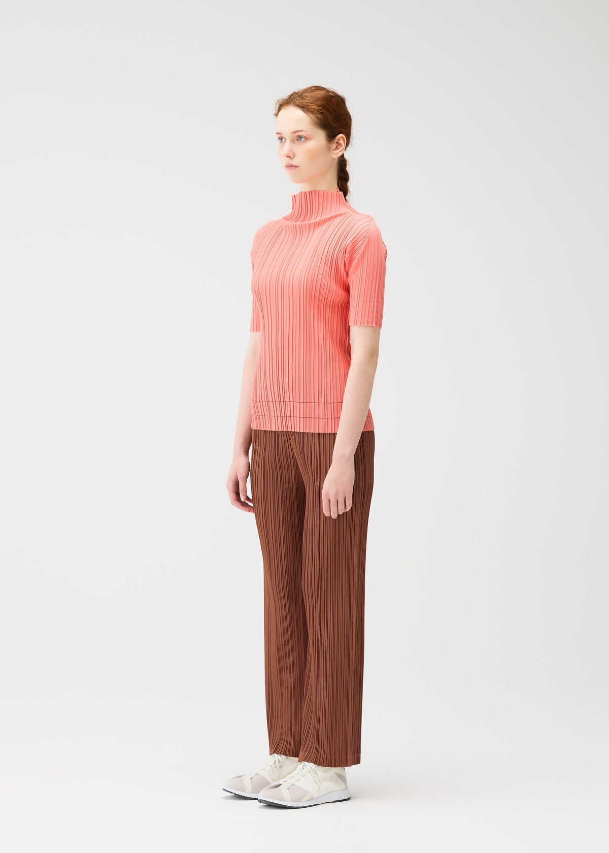 A woman with red braided hair stands against a white background, wearing the SOFT PLEATS brown pleated pants by PLEATS PLEASE ISSEY MIYAKE—styled with a pink top and white sneakers, facing slightly left and looking forward. Perfect for spring. view 6 of 7