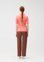 A person with long brown hair in a braid faces away, wearing SOFT PLEATS pants by PLEATS PLEASE ISSEY MIYAKE, a ribbed pink top, and white shoes—an effortlessly stylish spring look against a plain light background.