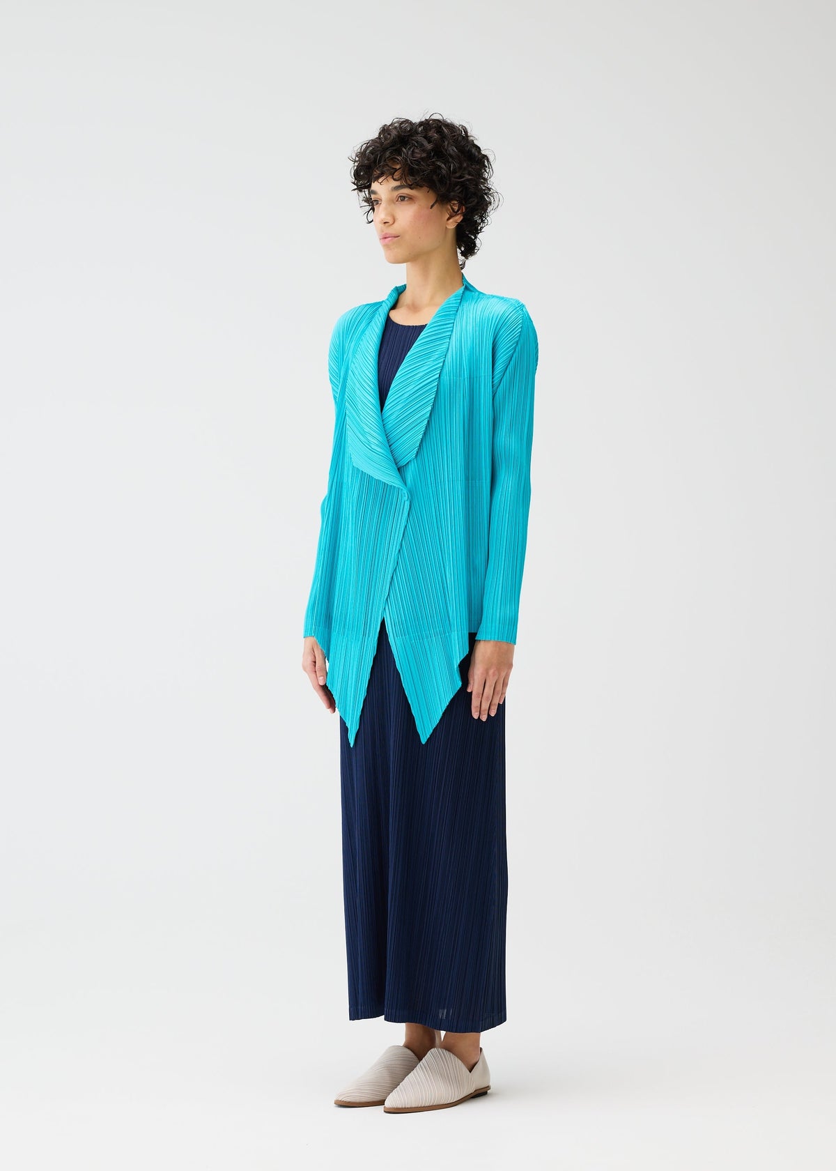 A person with short curly hair stands against a plain background, wearing the MONTHLY COLORS : JANUARY pleated cardigan from PLEATS PLEASE ISSEY MIYAKE over a long navy dress and light beige slip-on shoes. view 8 of 9