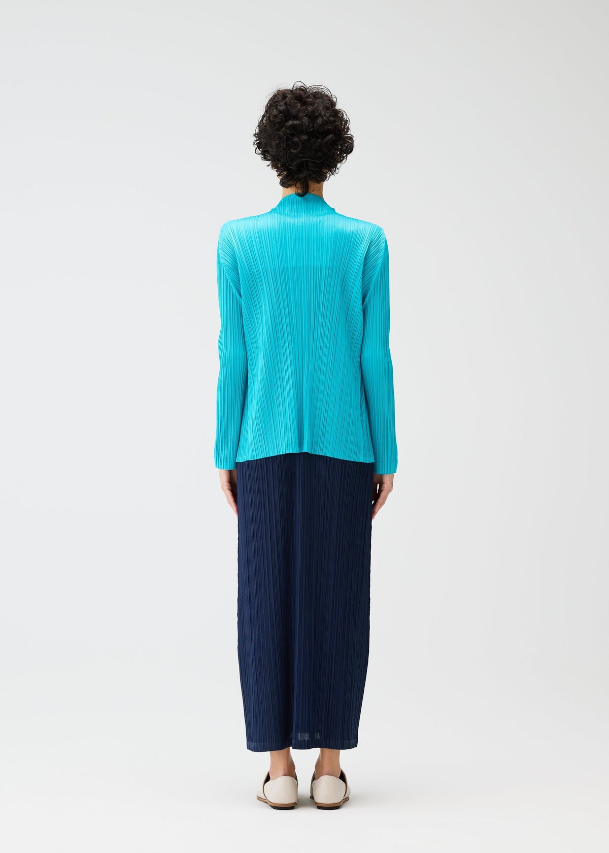 A person with short curly hair, seen from behind, wears the PLEATS PLEASE ISSEY MIYAKE MONTHLY COLORS: JANUARY collection—a bright turquoise cardigan and long navy pleated skirt—paired with white flats against a plain white background. view 9 of 9