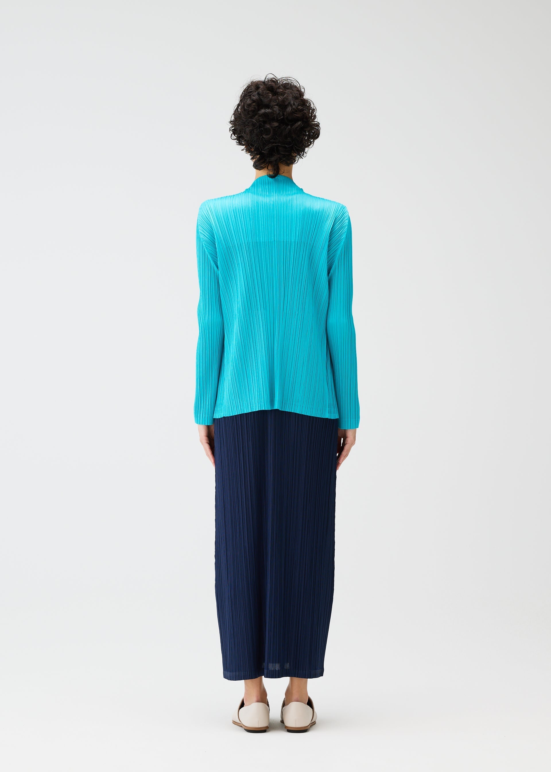 Zoomed view A person with short curly hair, seen from behind, wears the PLEATS PLEASE ISSEY MIYAKE MONTHLY COLORS: JANUARY collection—a bright turquoise cardigan and long navy pleated skirt—paired with white flats against a plain white background.