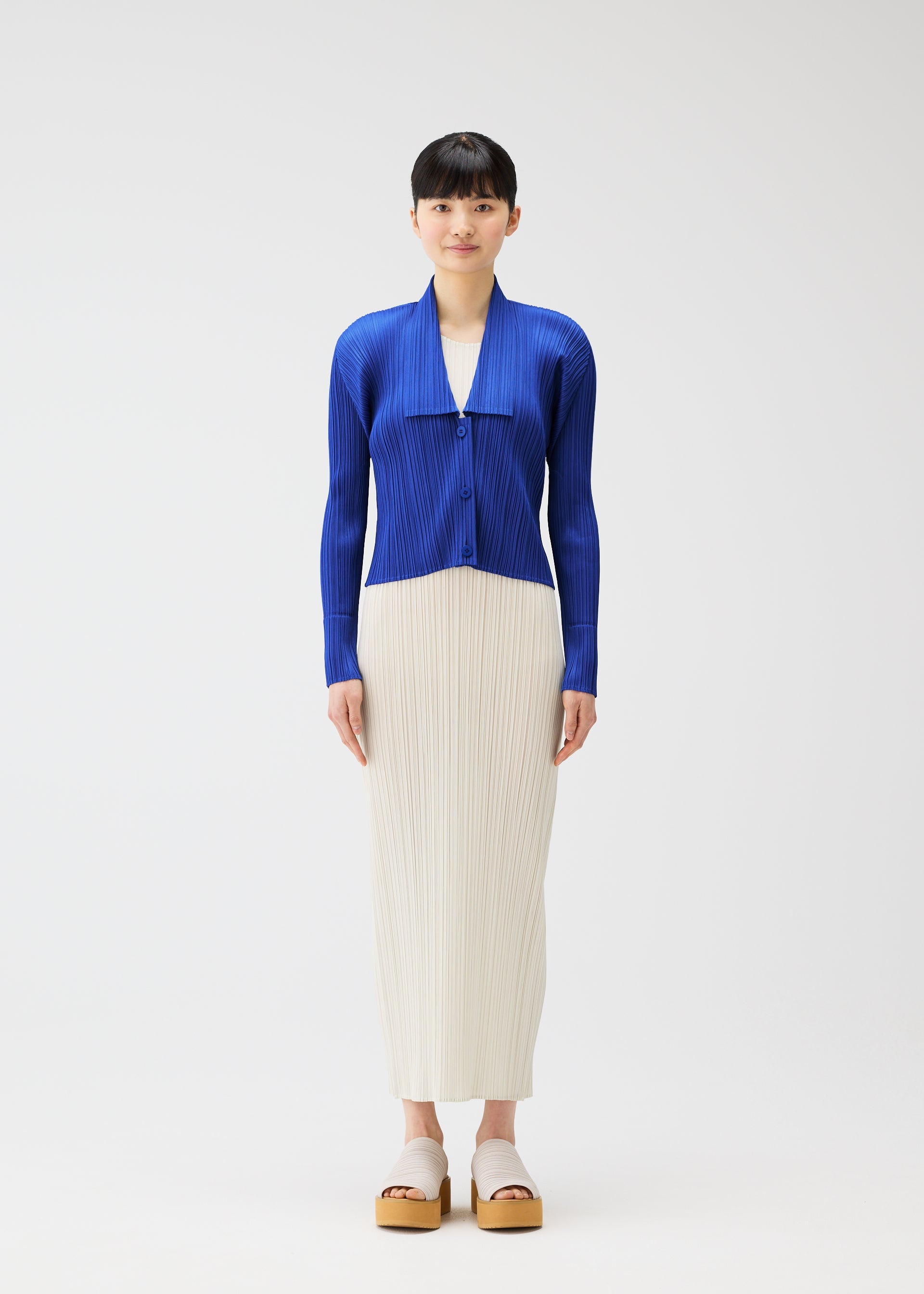 Zoomed view A person faces forward, wearing MONTHLY COLORS : MAY by PLEATS PLEASE ISSEY MIYAKE—a bright blue collared cardigan with button closures over a white ankle-length pleated skirt and beige platform sandals, set against a plain white background.