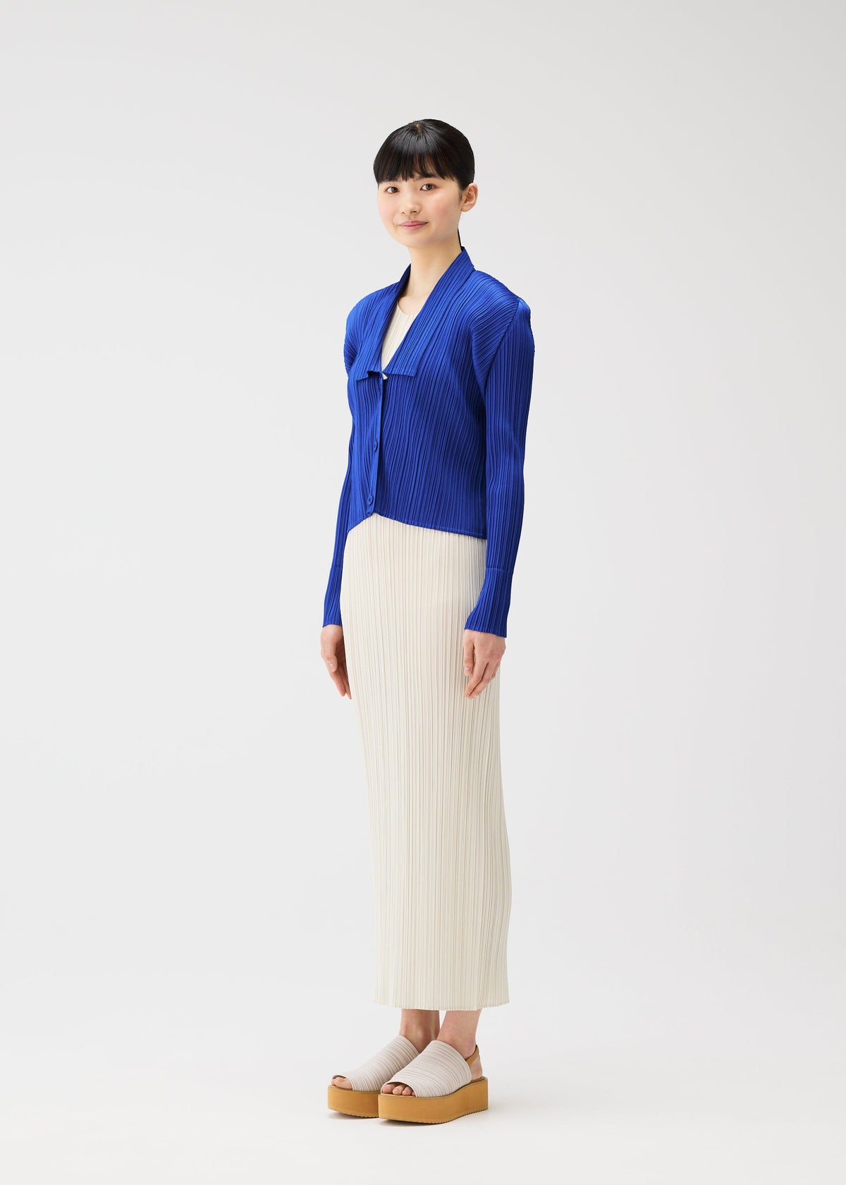 A woman with straight black hair and bangs models the PLEATS PLEASE ISSEY MIYAKE MONTHLY COLORS : MAY royal blue collared cardigan, paired with a cream skirt and beige platform sandals, smiling gently in a white studio setting. view 8 of 9