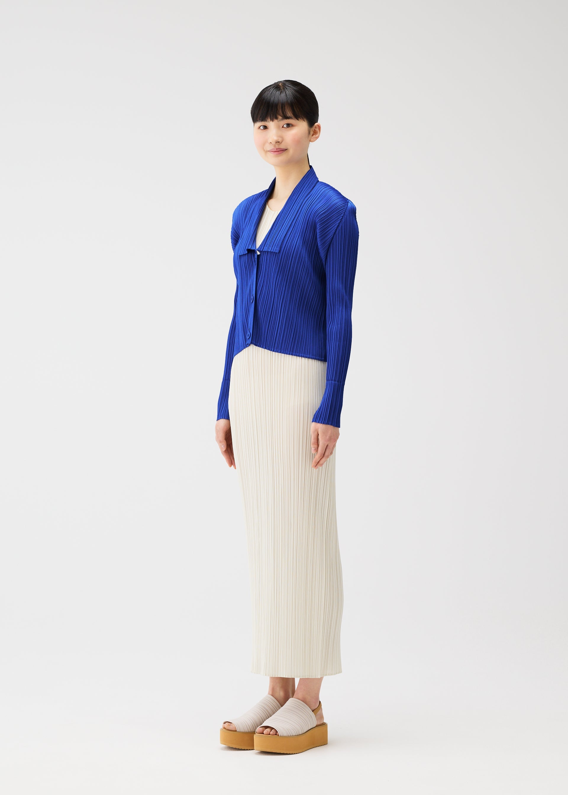 Zoomed view A woman with straight black hair and bangs models the PLEATS PLEASE ISSEY MIYAKE MONTHLY COLORS : MAY royal blue collared cardigan, paired with a cream skirt and beige platform sandals, smiling gently in a white studio setting.