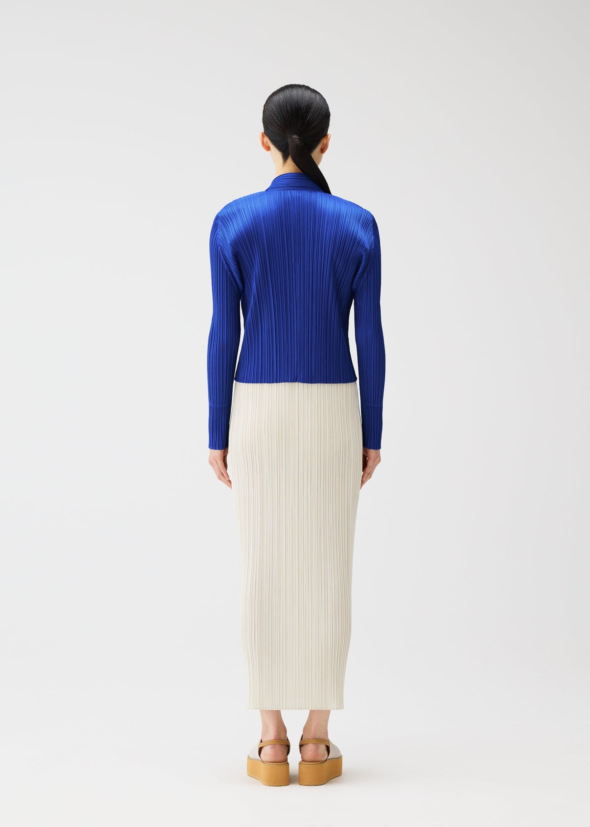 A person faces away, dressed in MONTHLY COLORS : MAY by PLEATS PLEASE ISSEY MIYAKE—a royal blue pleated top with soft gradation, an off-white pleated skirt, and platform sandals. Their long dark hair is tied back against a white background. view 9 of 9