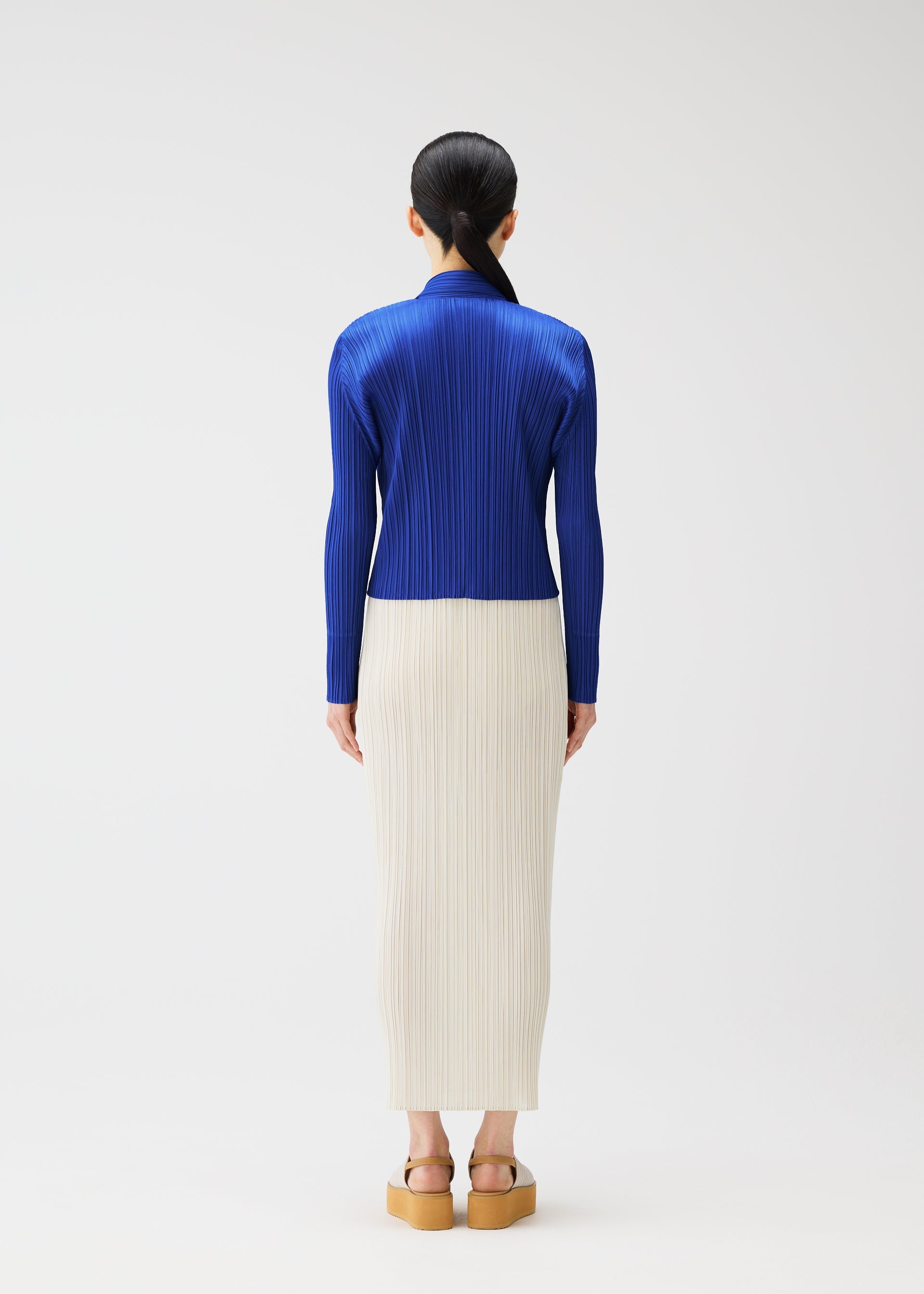 Zoomed view A person faces away, dressed in MONTHLY COLORS : MAY by PLEATS PLEASE ISSEY MIYAKE—a royal blue pleated top with soft gradation, an off-white pleated skirt, and platform sandals. Their long dark hair is tied back against a white background.