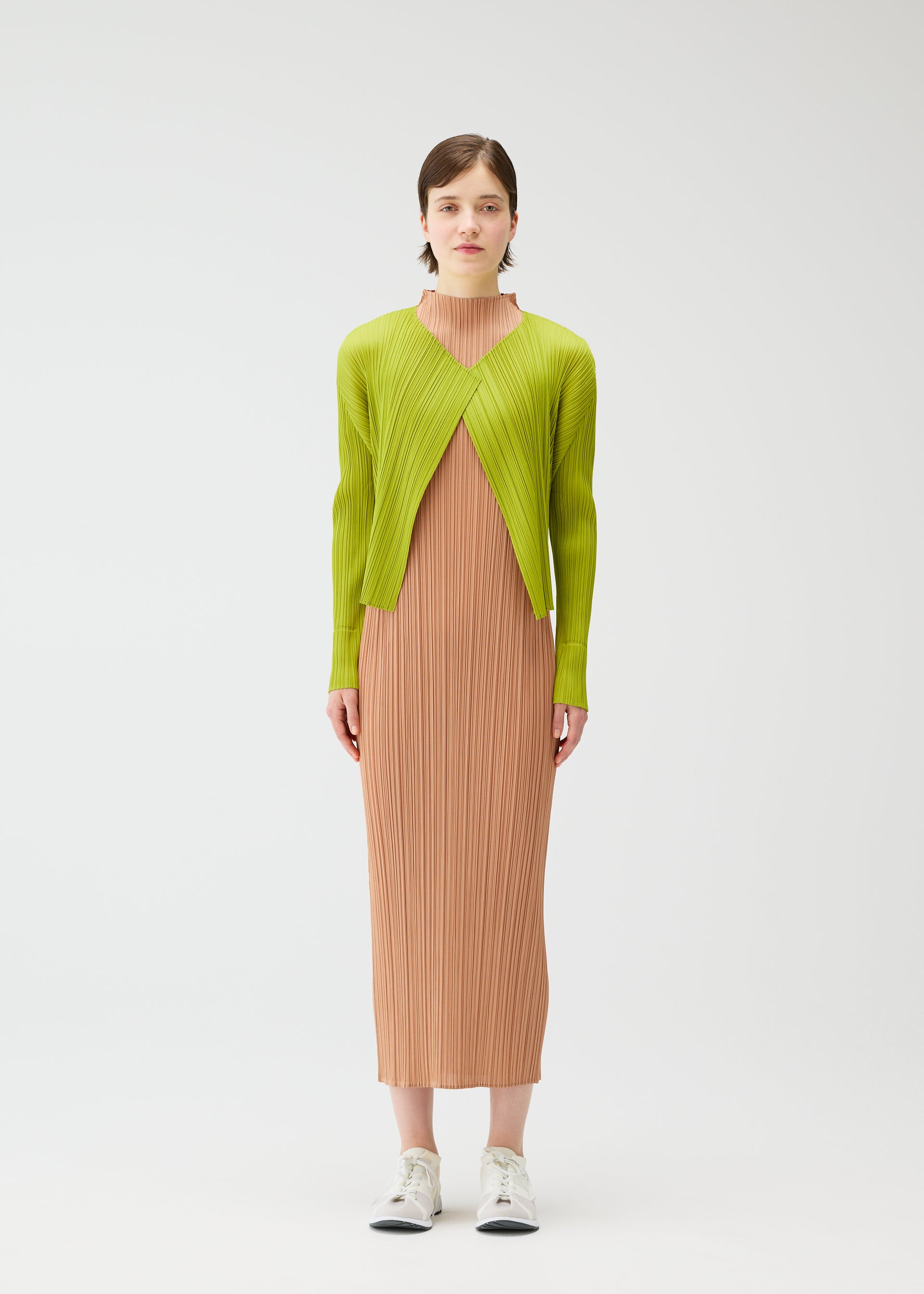 Zoomed view A woman faces forward in a neutral pose, wearing the MONTHLY COLORS : JUNE long light brown pleated dress by PLEATS PLEASE ISSEY MIYAKE, paired with a fruit-and-vegetable-inspired lime green cardigan and white sneakers against a white background.