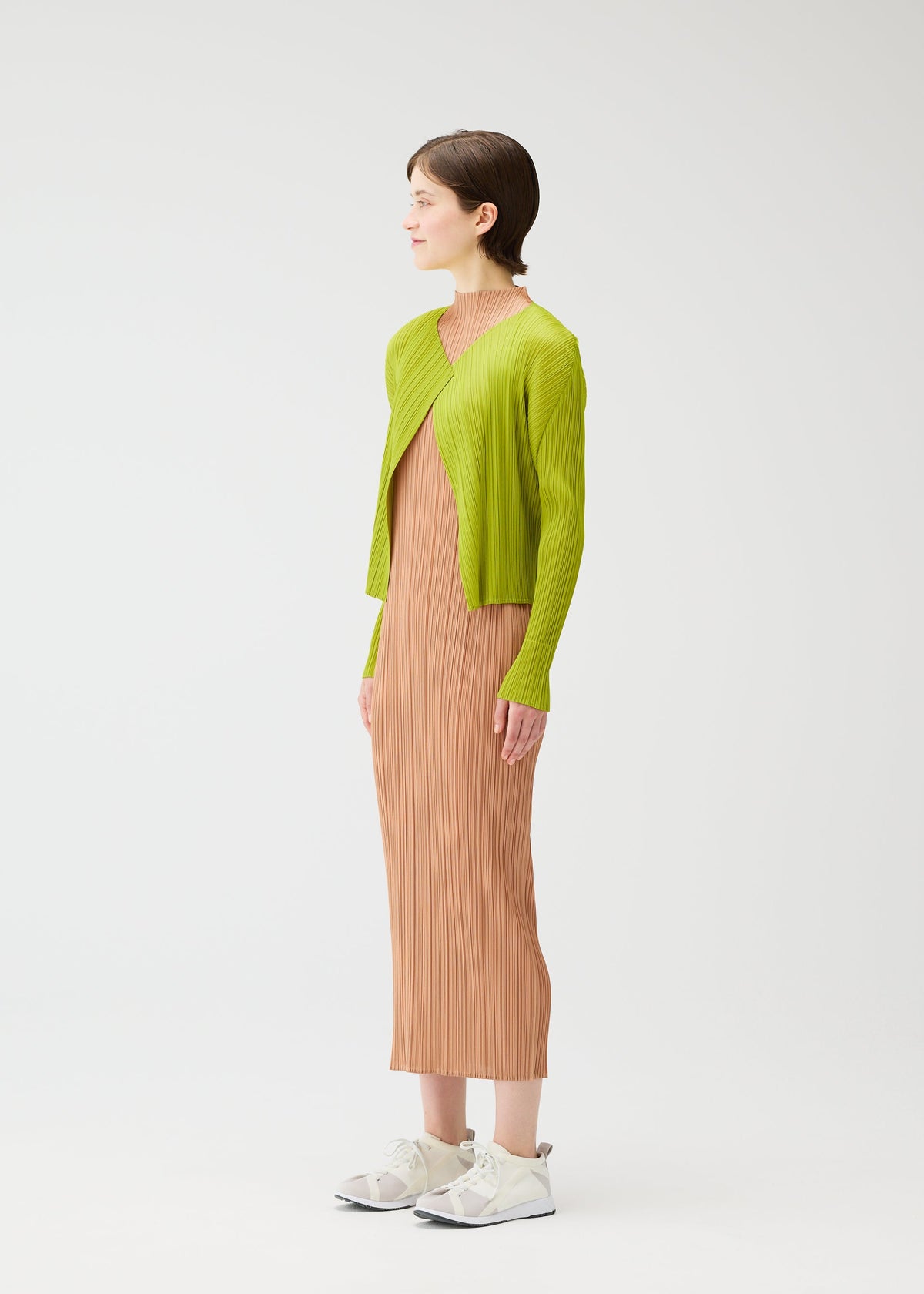 A woman with short brown hair and a neutral expression models MONTHLY COLORS : JUNE by PLEATS PLEASE ISSEY MIYAKE—a long beige pleated dress paired with a lime green cardigan and white sneakers, standing sideways against a plain background. view 8 of 9