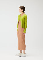 A woman with short brown hair and a neutral expression models MONTHLY COLORS : JUNE by PLEATS PLEASE ISSEY MIYAKE—a long beige pleated dress paired with a lime green cardigan and white sneakers, standing sideways against a plain background.